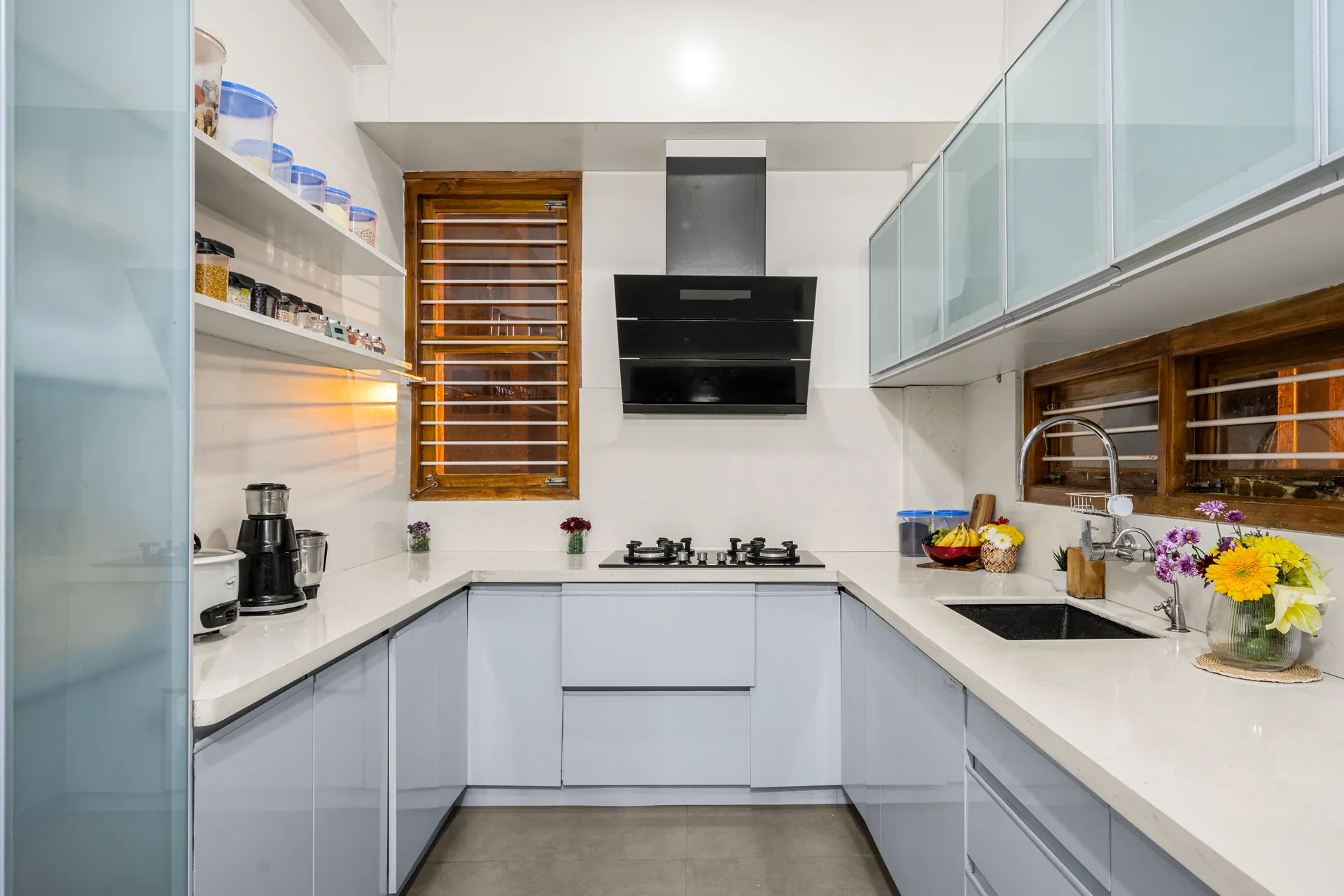 Galley-style kitchen with white counters, frosted glass wall cabinets, black hood and gas hob, and a sink under a wooden window.