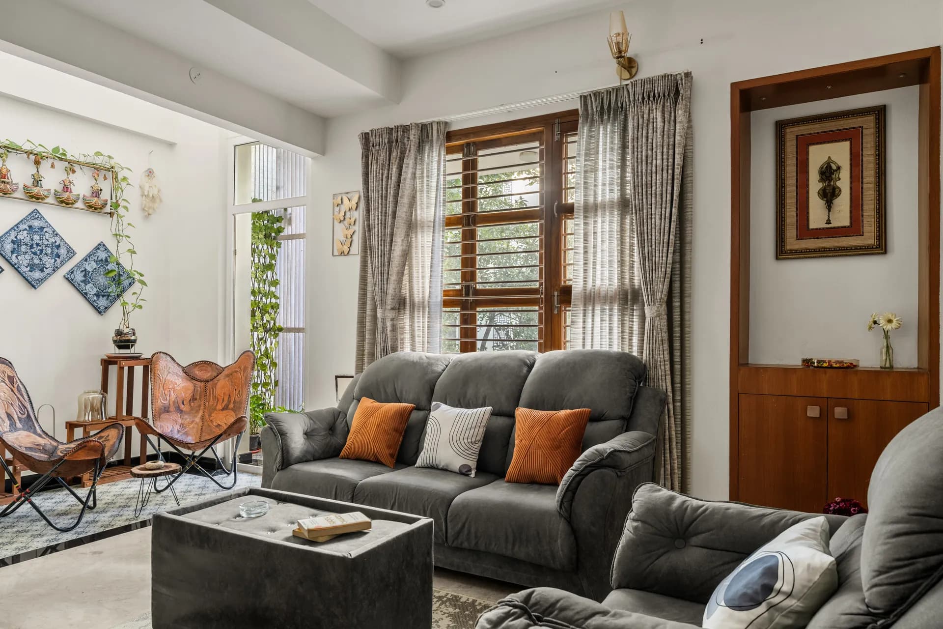 Living room with grey sofa, orange and patterned cushions, leather butterfly chairs and wooden-shuttered window with curtains.