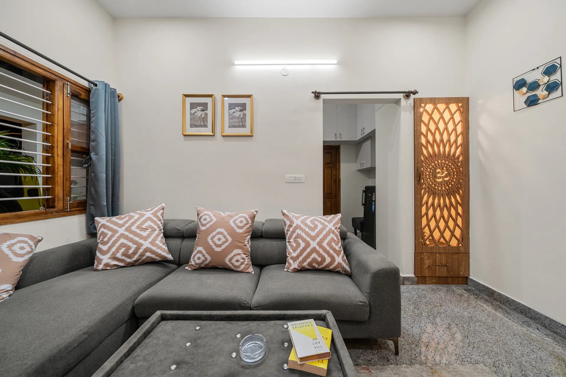 Living room with grey sectional sofa, patterned cushions, a lit carved wooden cabinet and a coffee table with books.