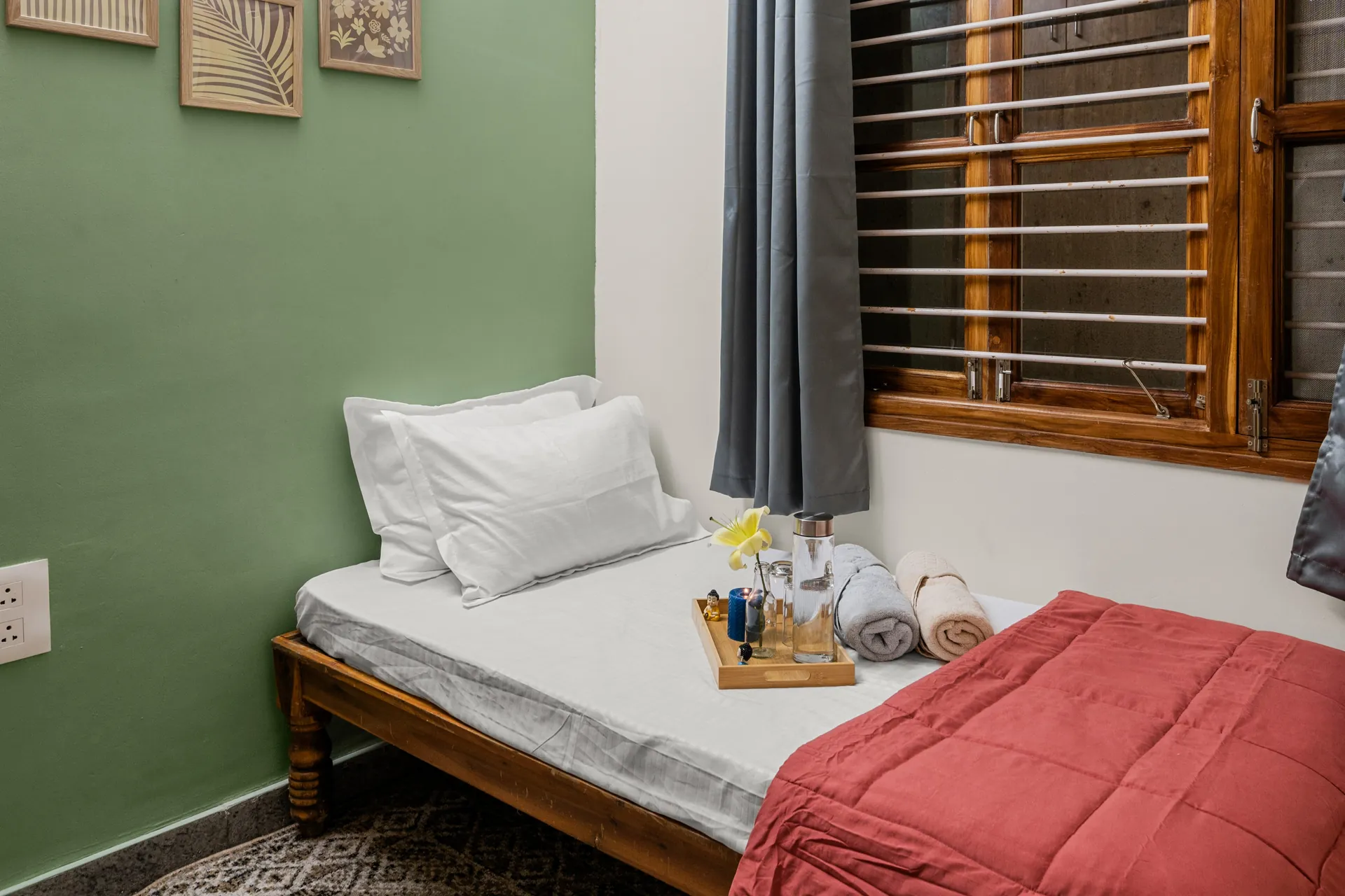 Small bedroom with a daybed, white pillows, a tray with a flower and water bottle, and folded towels by a wooden-shuttered window.