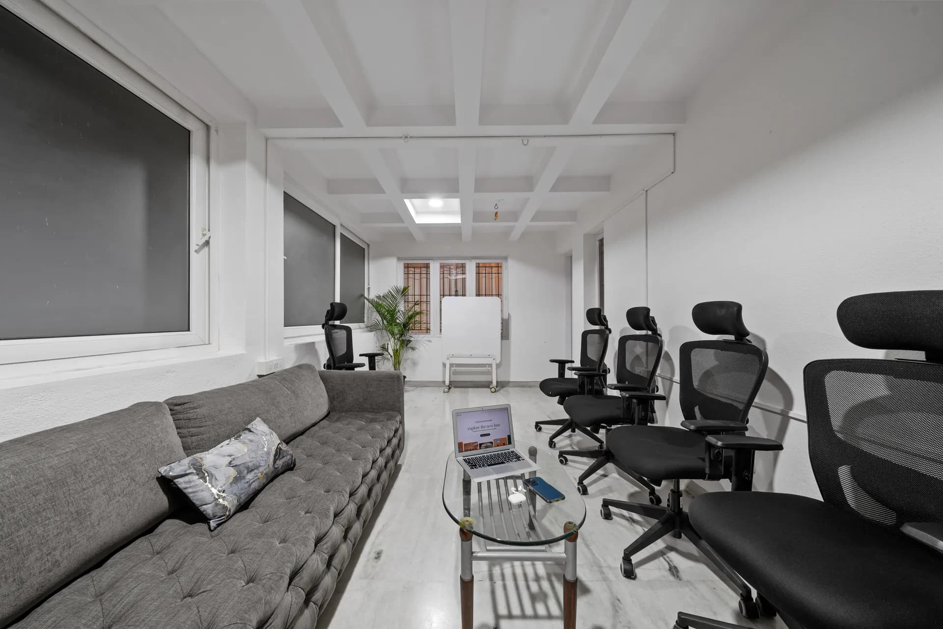 A bright workspace with a grey tufted sofa, four black mesh task chairs, and a glass coffee table with a laptop.
