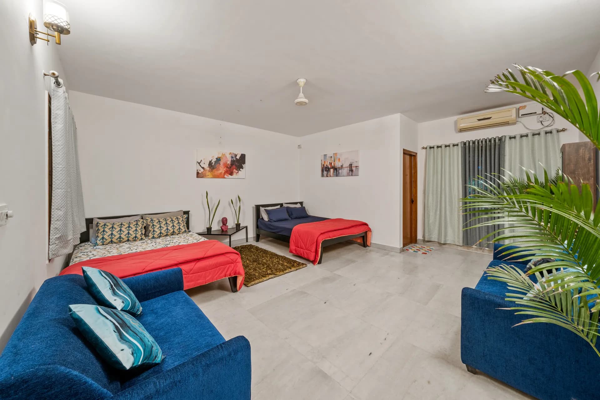 Bedroom with two beds, blue sofas, and a potted palm by the sliding curtains and wall-mounted air conditioner.