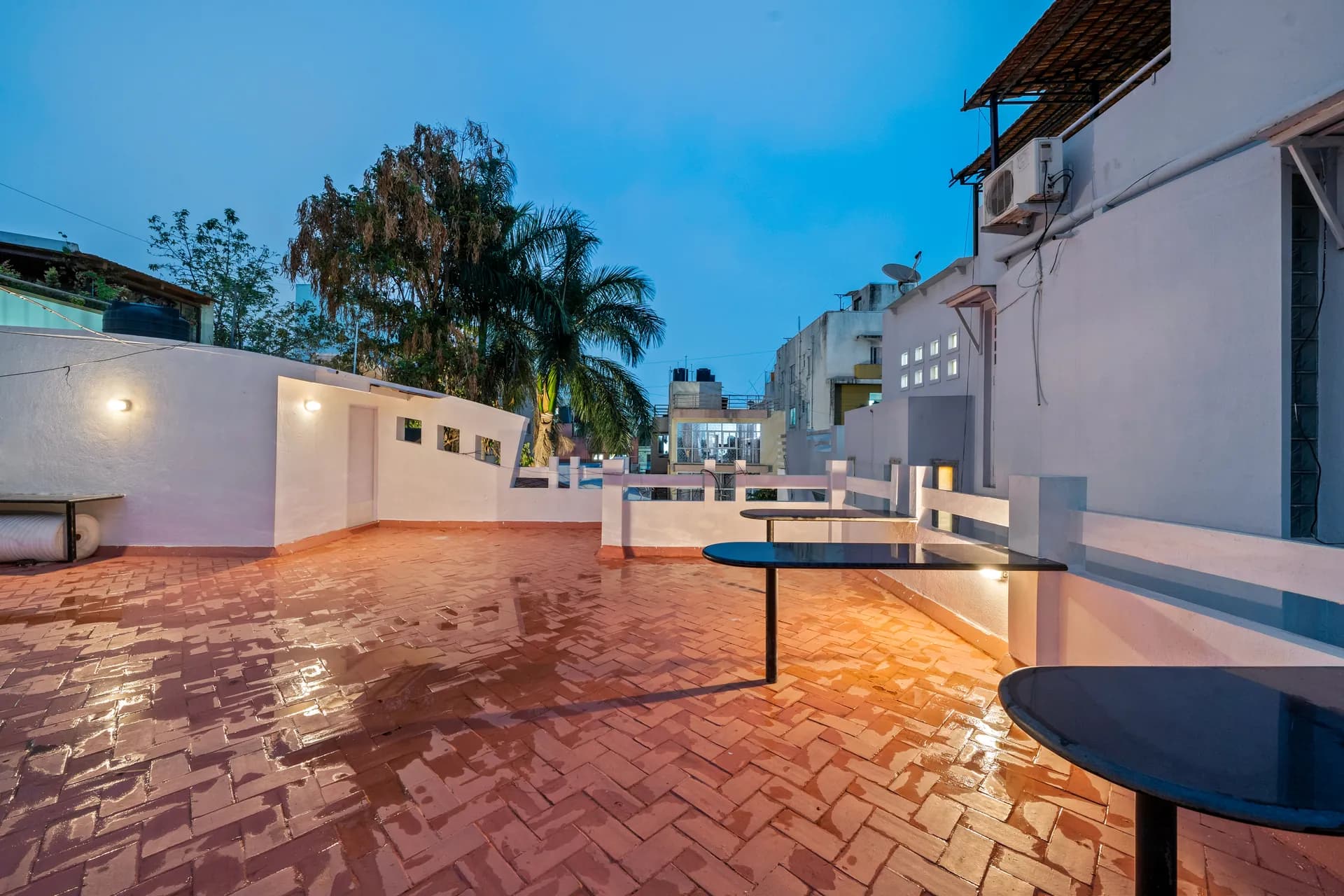 Open brick-paved rooftop terrace at dusk with black round tables, white walls and a palm tree beyond.