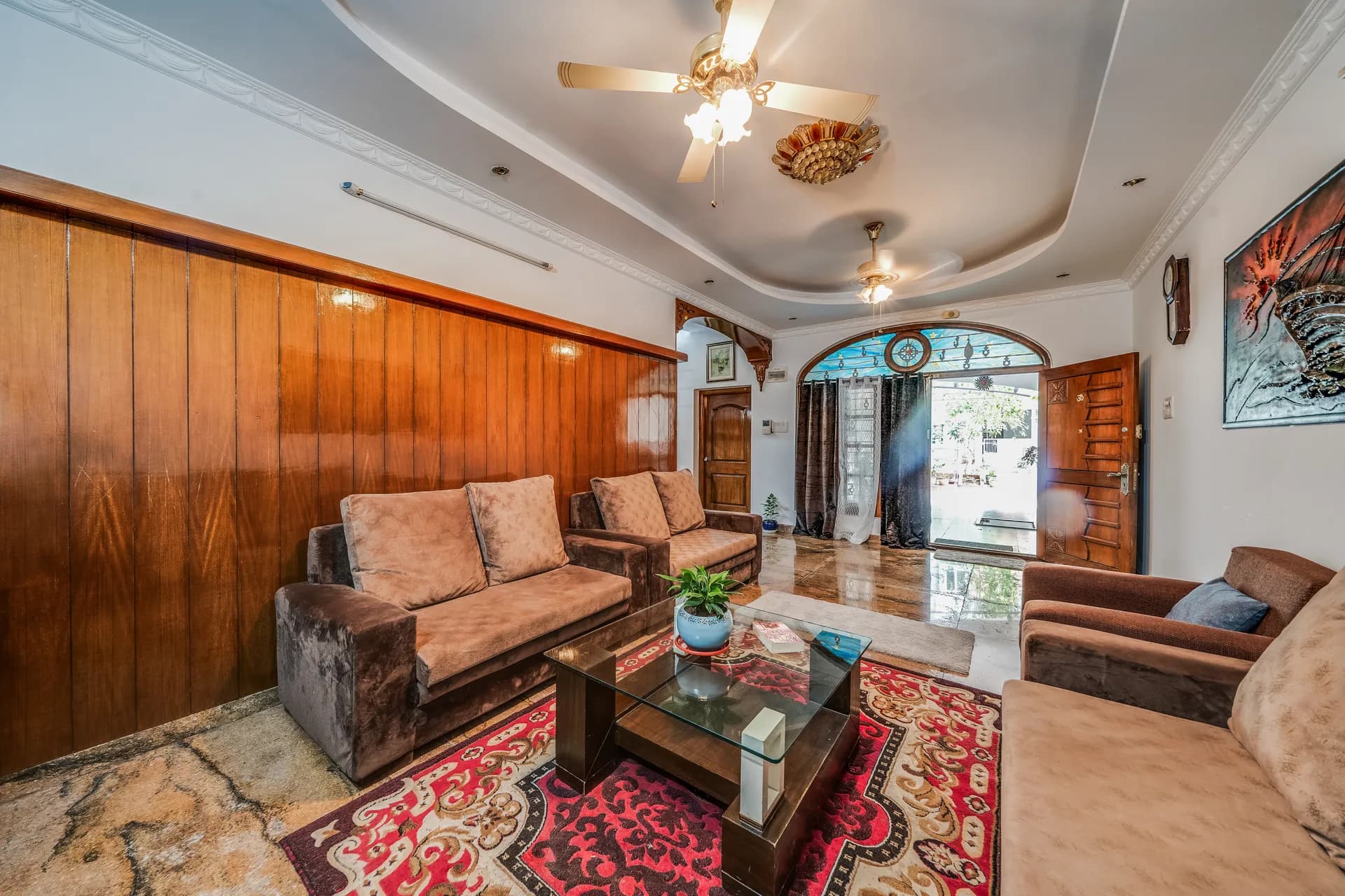 Living room with wood-panelled wall, brown sofas, glass coffee table and red patterned rug, opening to a courtyard.