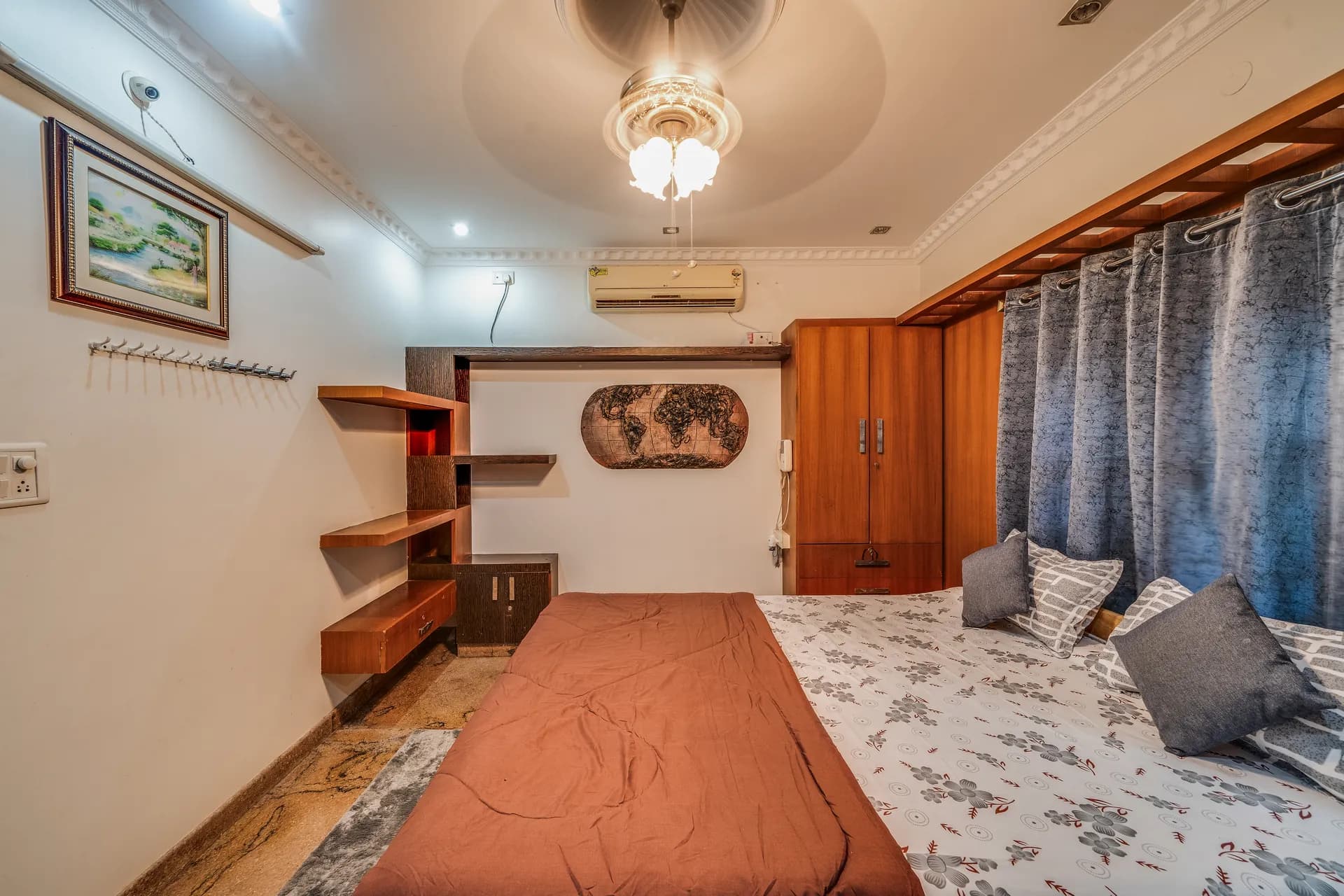 Bedroom with a ceiling fan, wall-mounted air conditioner, a double bed with grey patterned linen, and a wooden wardrobe.