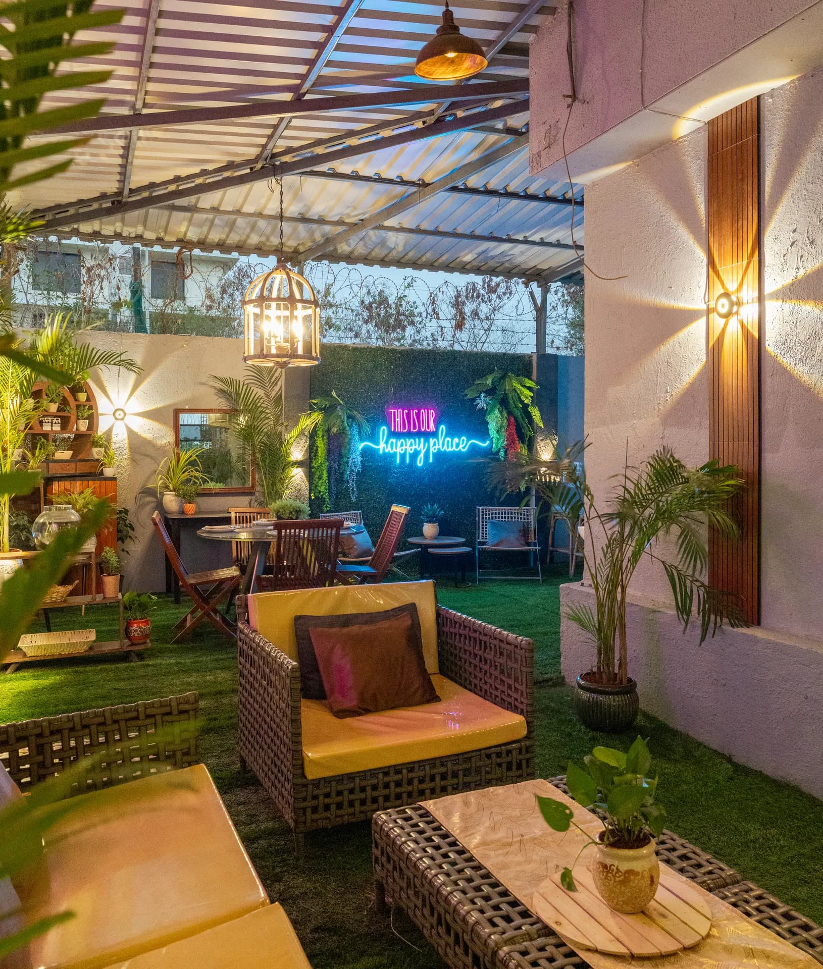 Covered garden terrace with wicker lounge seating, a neon 'This is our happy place' sign, and potted palms.