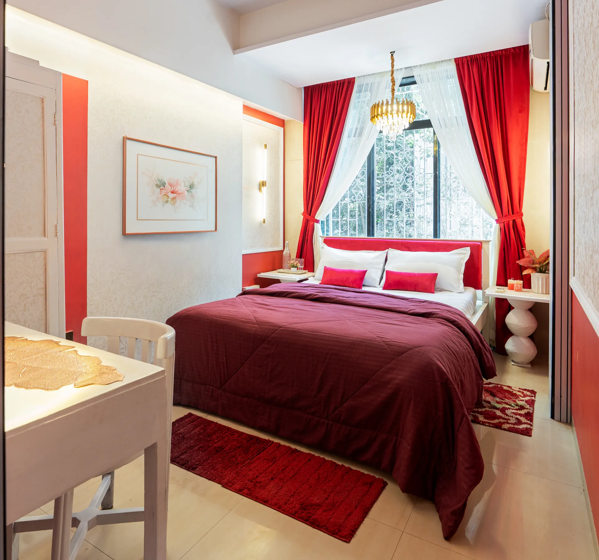 Bedroom with a made-up king bed in deep red, red curtains, and a gold chandelier over the window.