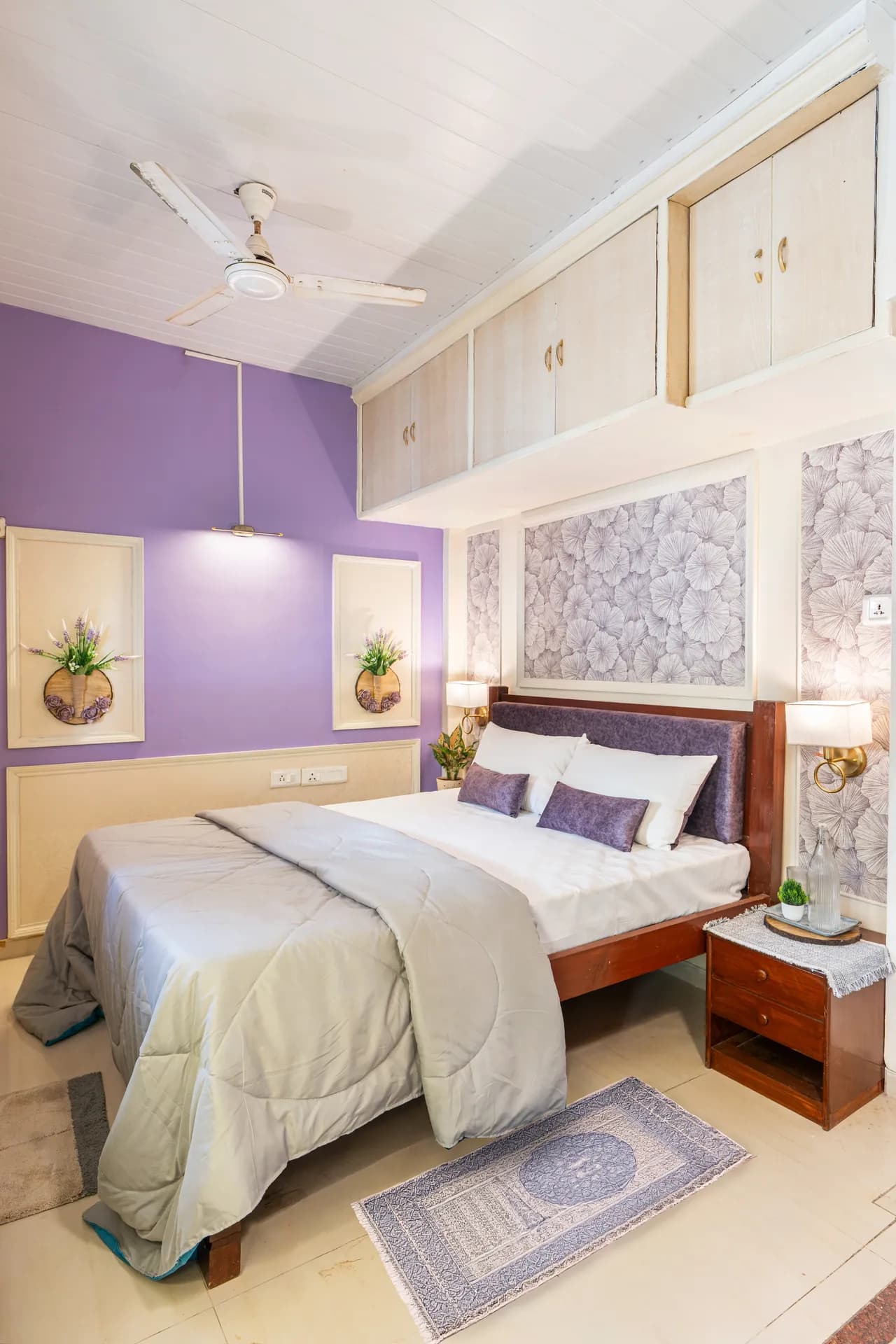 Bedroom with purple accent wall, queen bed with grey comforter, overhead cabinets and wall art.