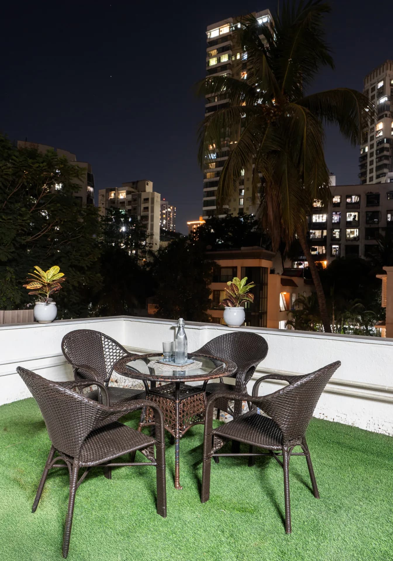Rooftop terrace with four wicker chairs and a glass table, artificial turf floor, palm tree and city towers at night.