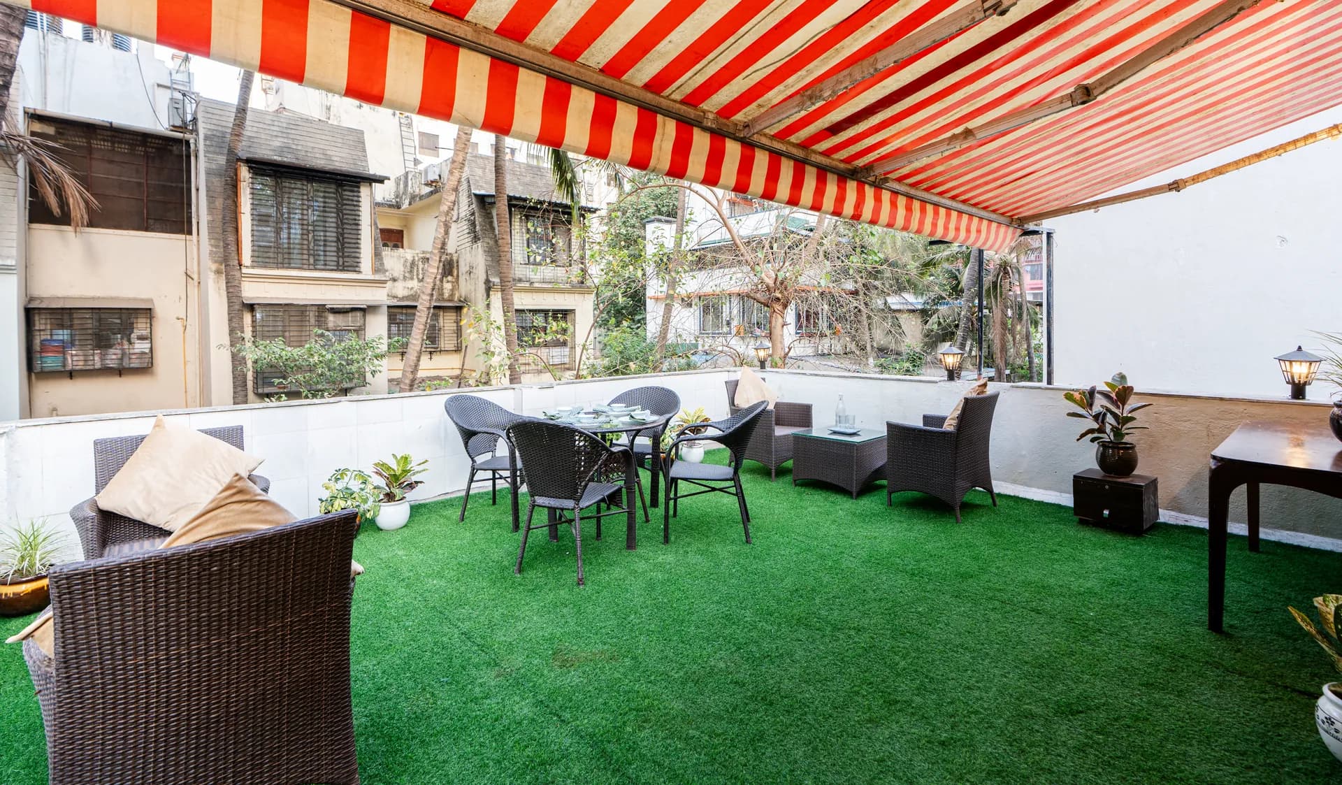 Covered terrace with artificial lawn, wicker lounge chairs, dining table and a striped awning overhead.