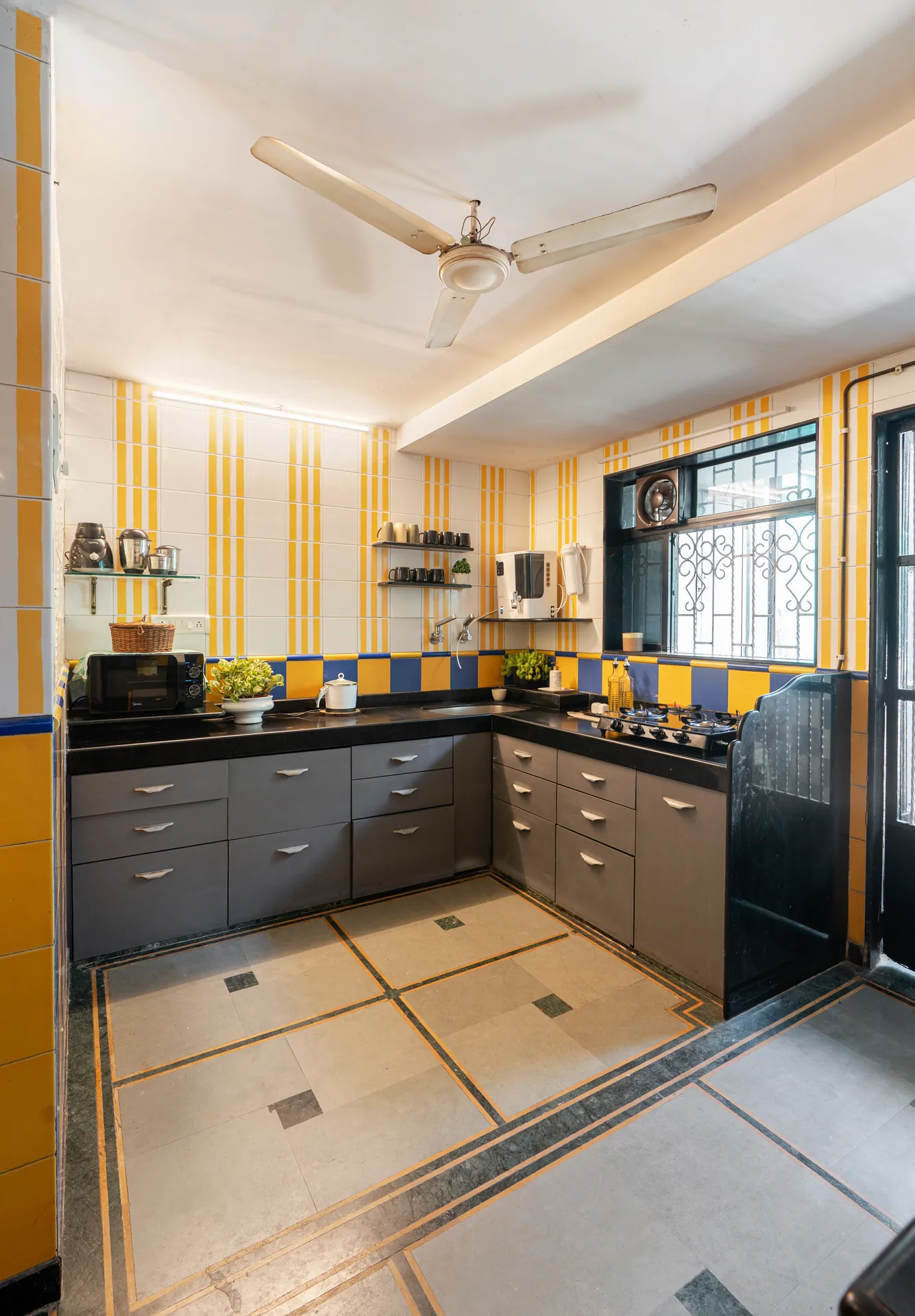 Galley-style kitchen with yellow and blue tiled walls, black worktop, grey cabinets, ceiling fan and gas hob.