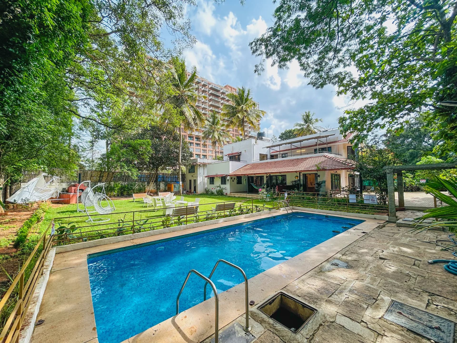 Outdoor pool with stone deck and lawn, backed by a tiled-roof villa and tall trees.
