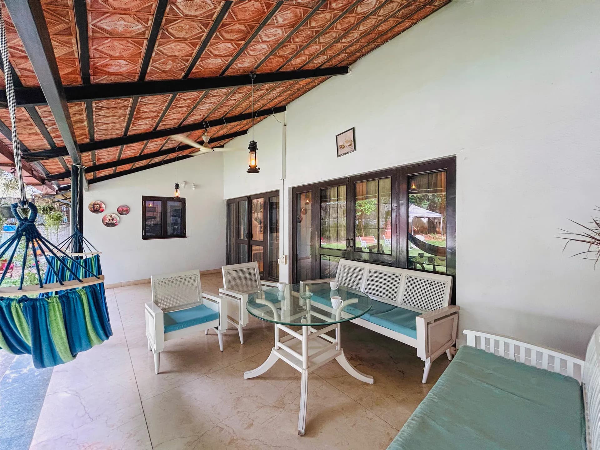 Covered patio with white cane seating, a glass dining table, a hanging hammock chair, and a terracotta tiled ceiling.
