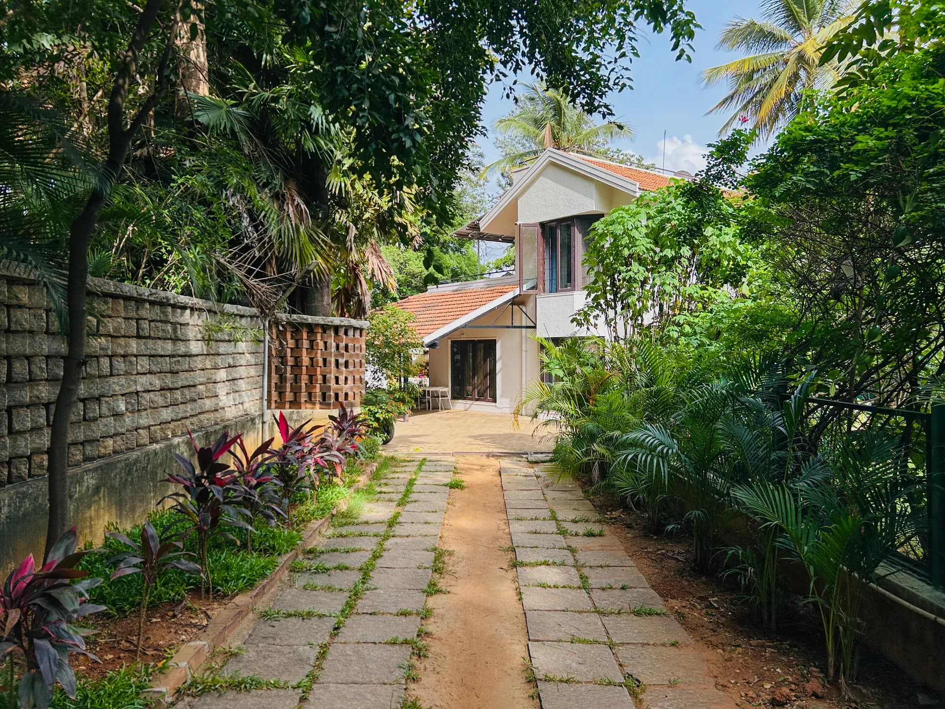 Paved path leads past lush palms and a red-tiled two-storey villa with a wooden-shuttered entrance.