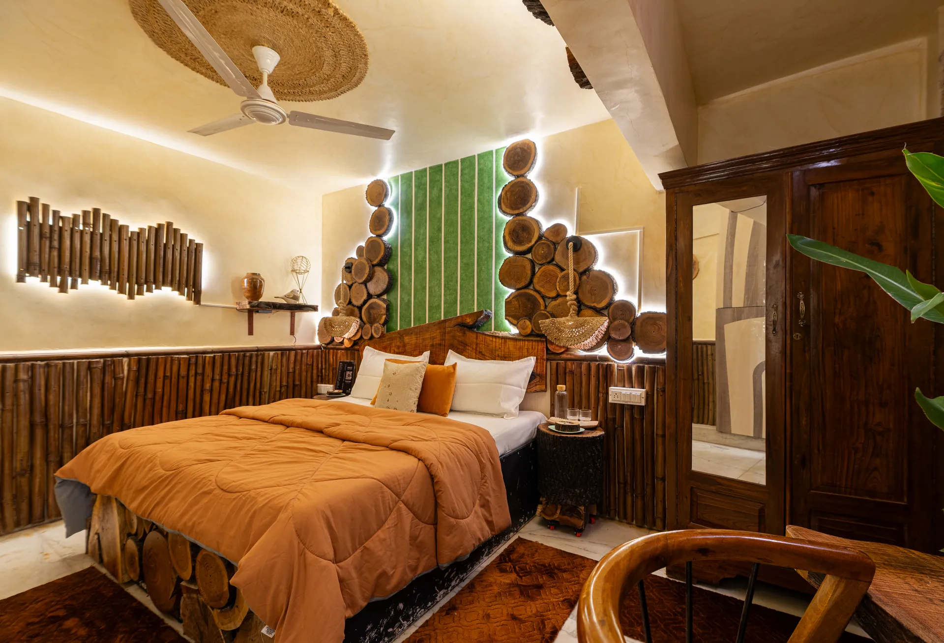 Bedroom with a king bed under a rattan ceiling fan, bamboo-panelled walls, and a wood-slice headboard backlit in green.