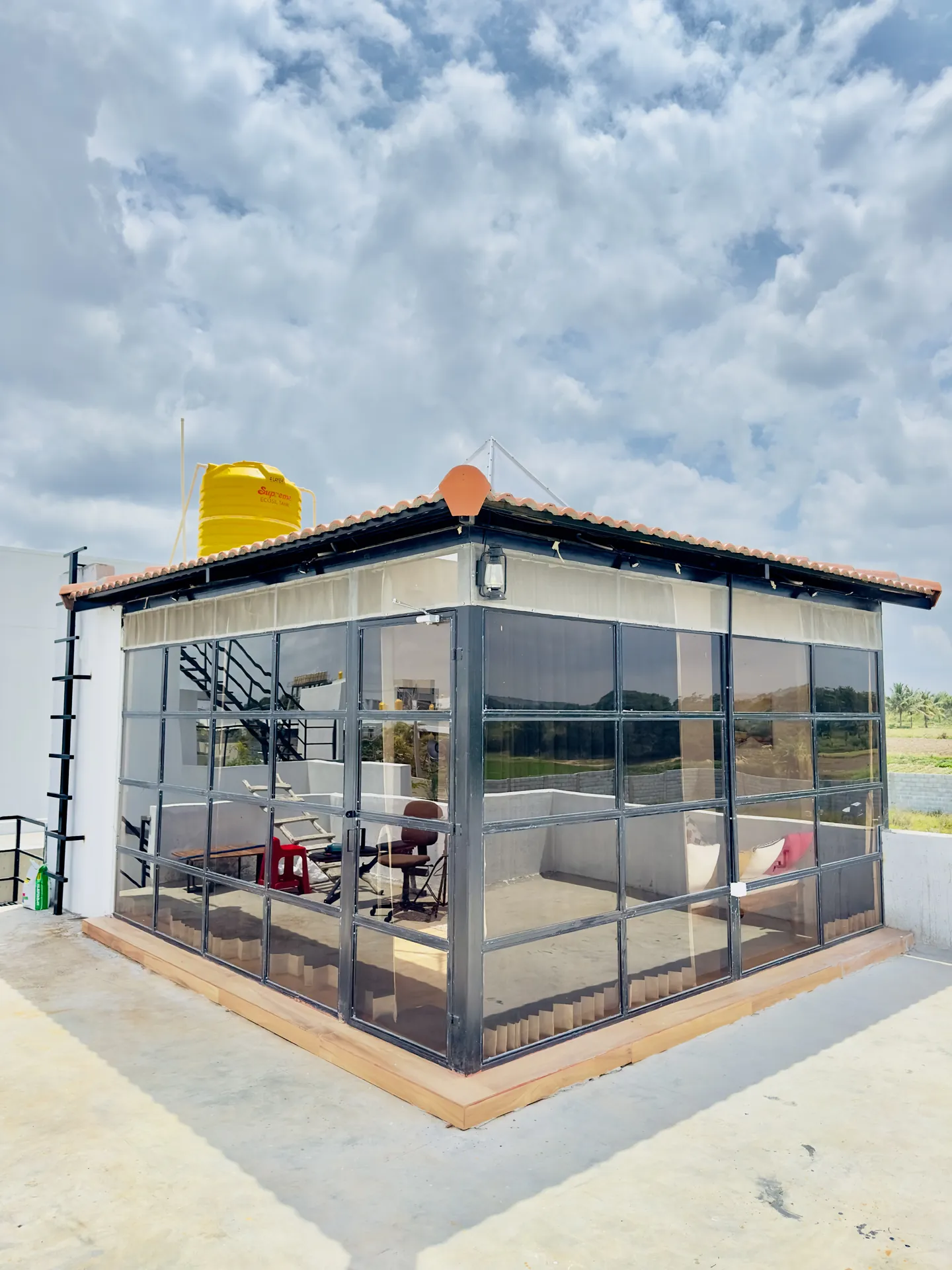 Glass-walled rooftop pavilion with a red-tiled roof, cushioned seating and a desk, beside a yellow water tank.