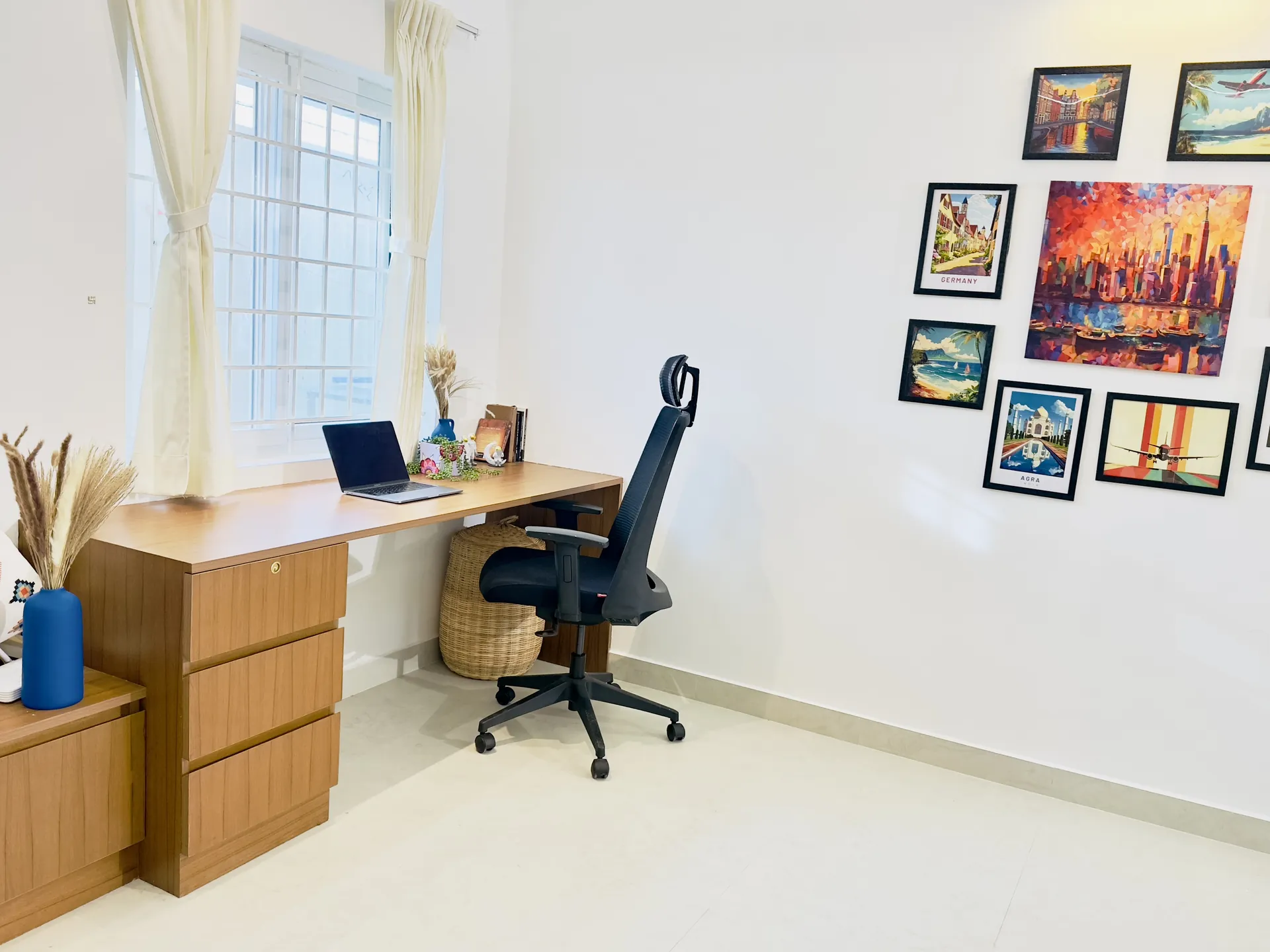 A bright workspace corner with a wooden desk, black ergonomic chair, laptop, and framed travel prints on the wall.