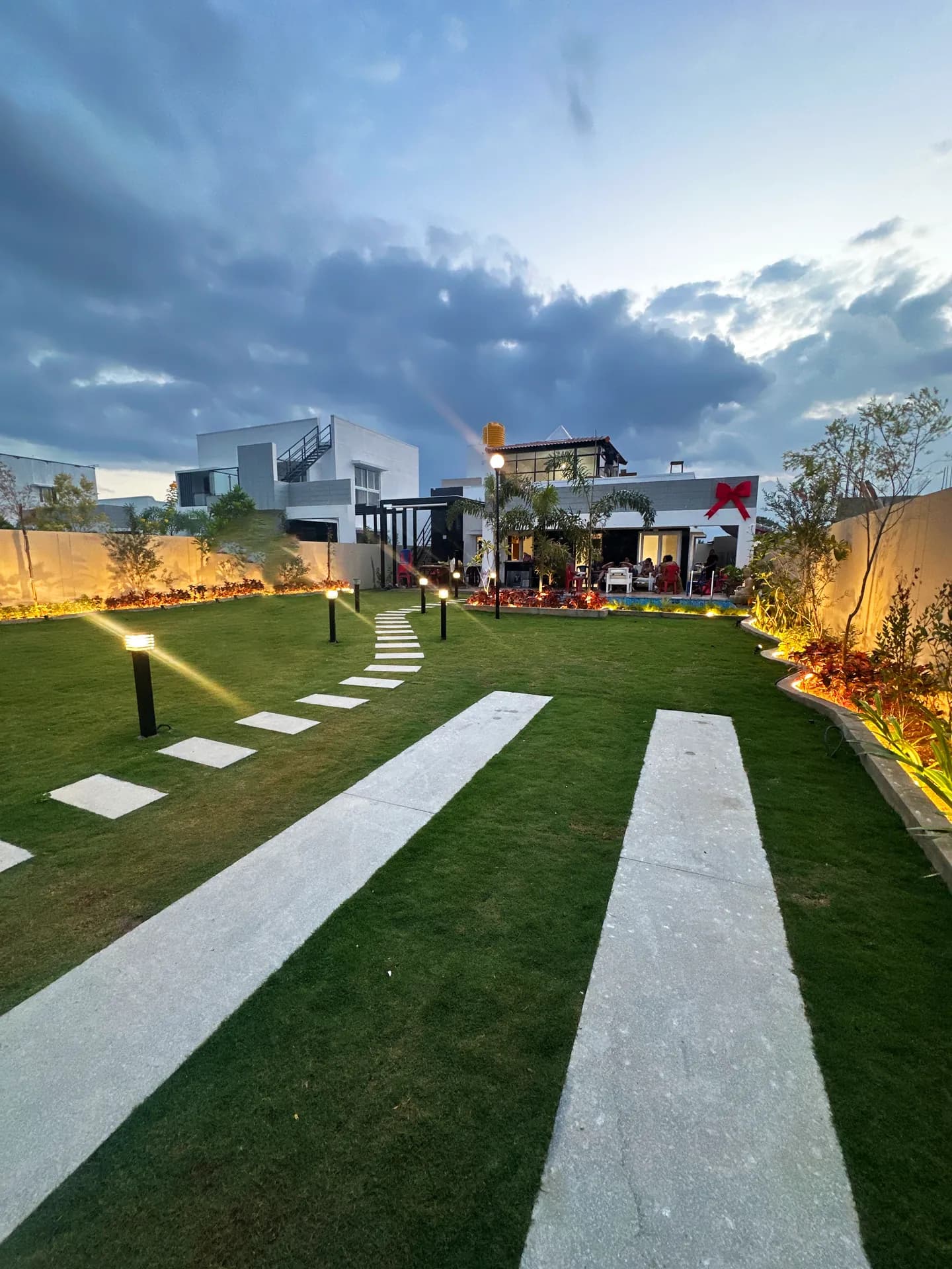 Evening view of a lit lawn garden with stepping-stone paths, bollard lights and a rear villa with a pool.