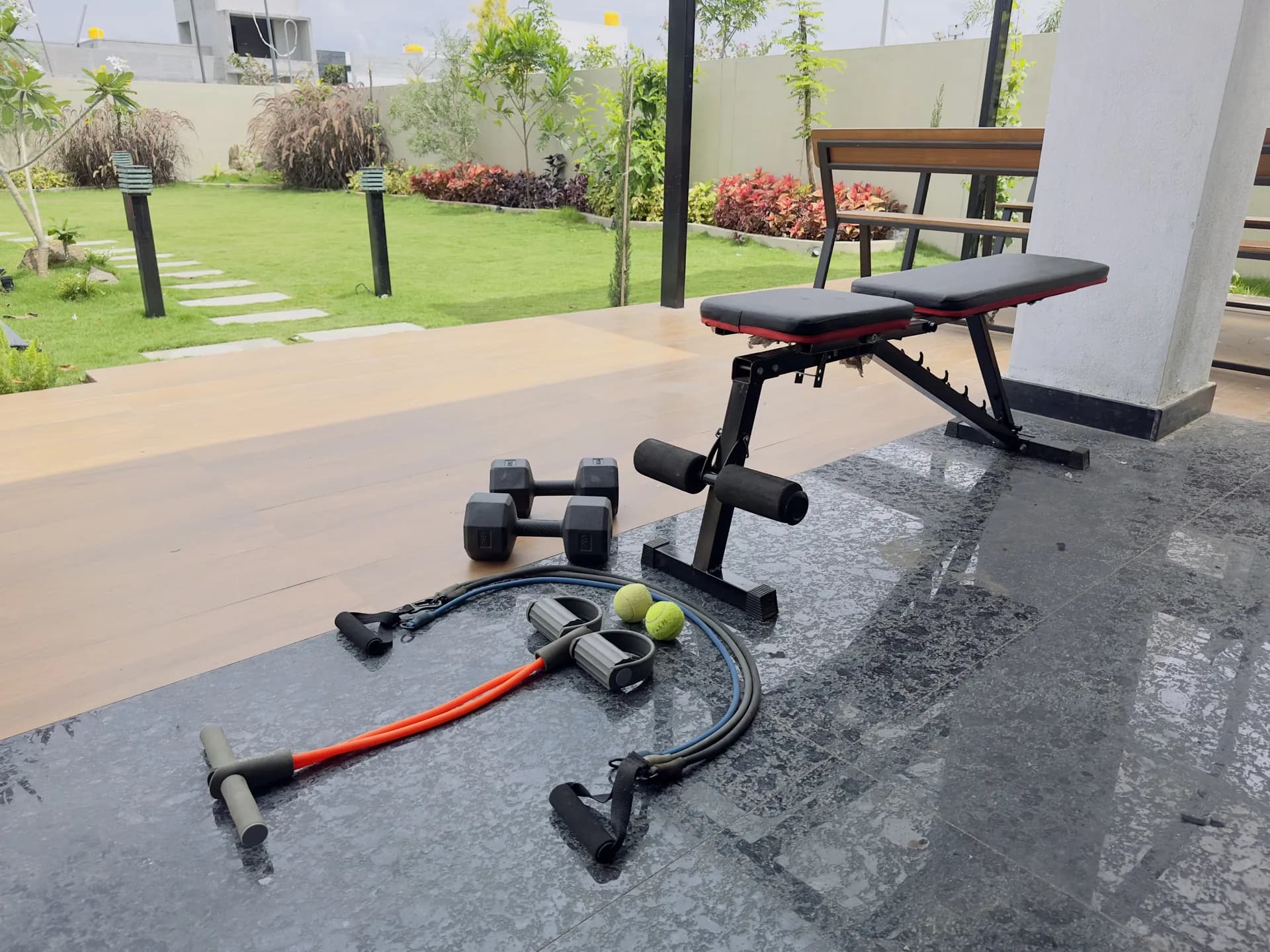 Outdoor gym corner with an adjustable weight bench, dumbbells, resistance bands and tennis balls beside a lawn.