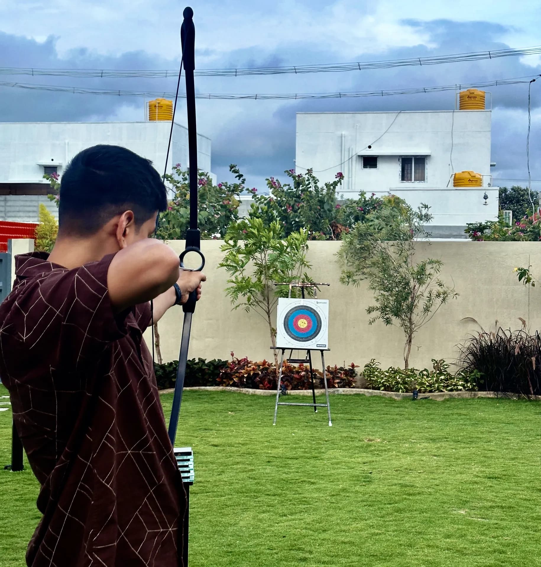 A man draws a recurve bow on a lawn facing an archery target against a white boundary wall.