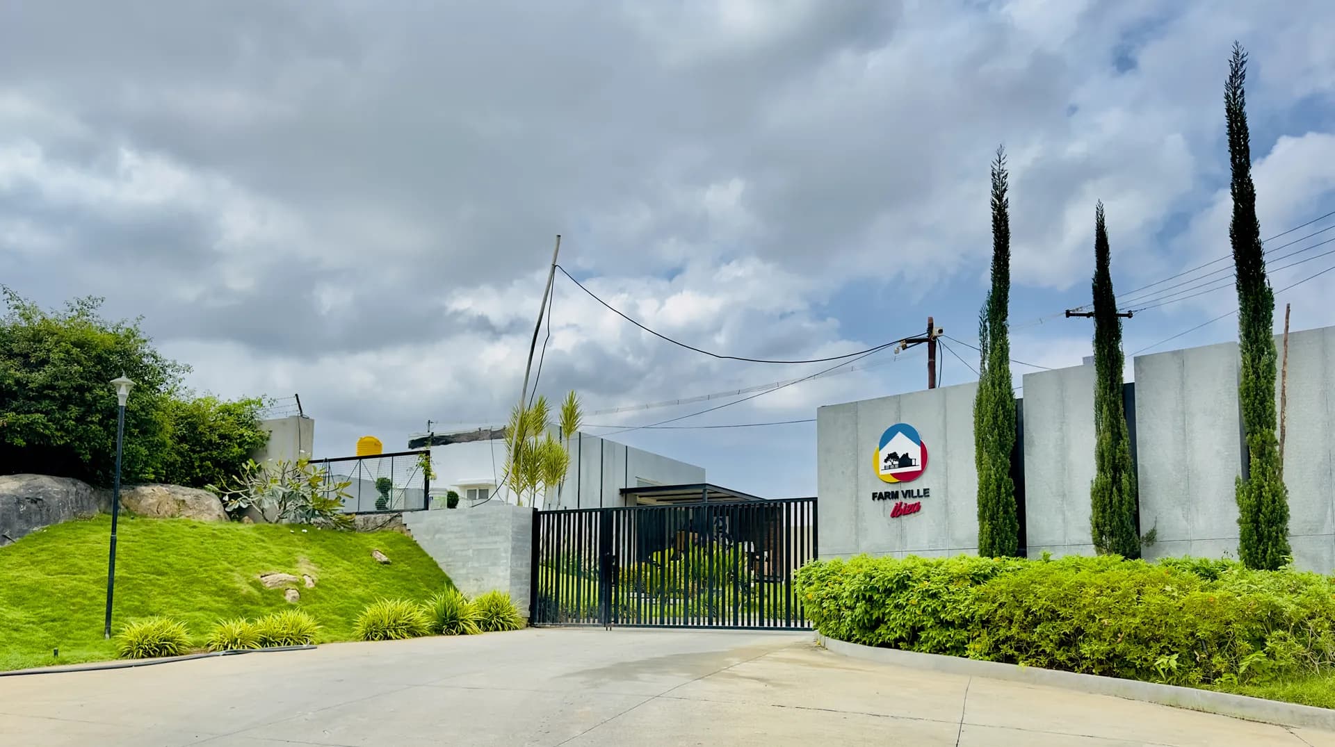 Gated villa entrance with a concrete wall, the Farmville Soul Club sign, cypress trees and a grassy slope.