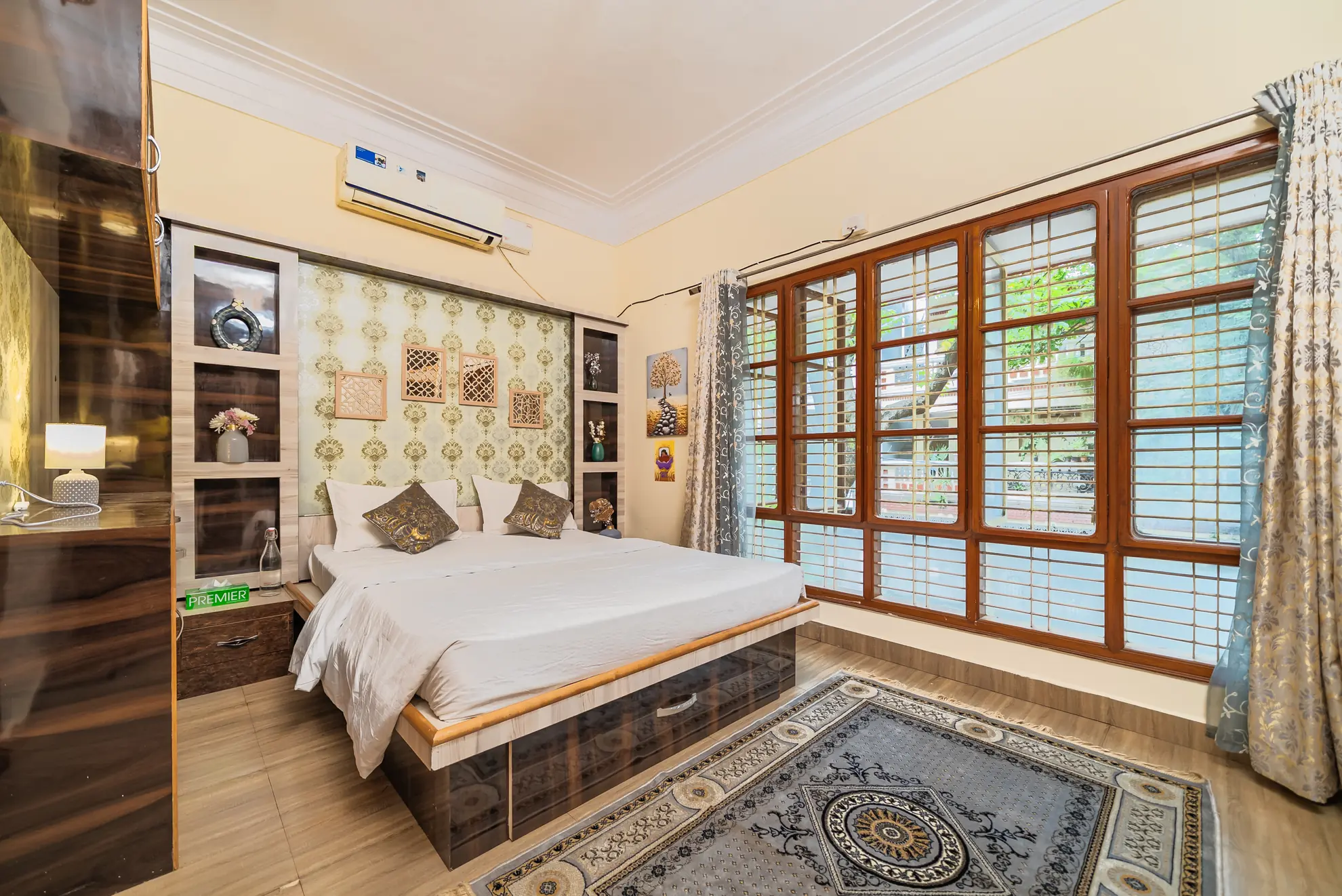 Bedroom with a large bed, wall-mounted air conditioner, and wide wooden-framed windows letting in daylight.