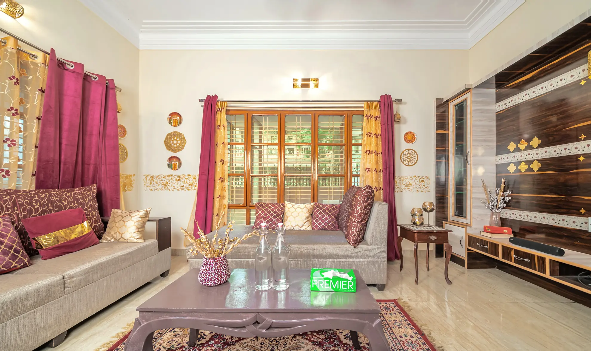 Living room with plum curtains, patterned sofas, a wooden-framed window and a low coffee table on a rug.