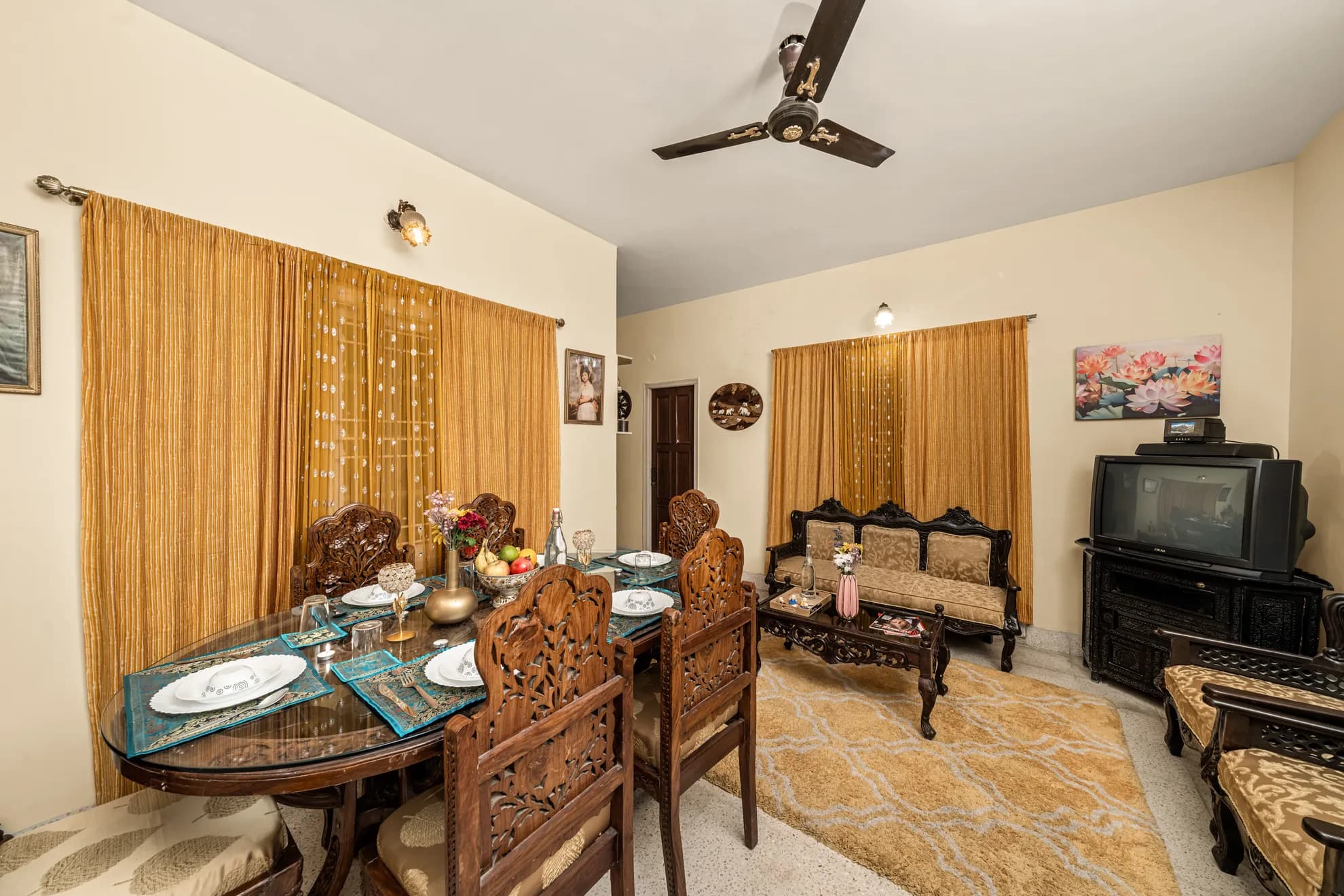 Dining area with a glass-topped table set for dinner, next to a sitting area with a carved sofa and a CRT television.
