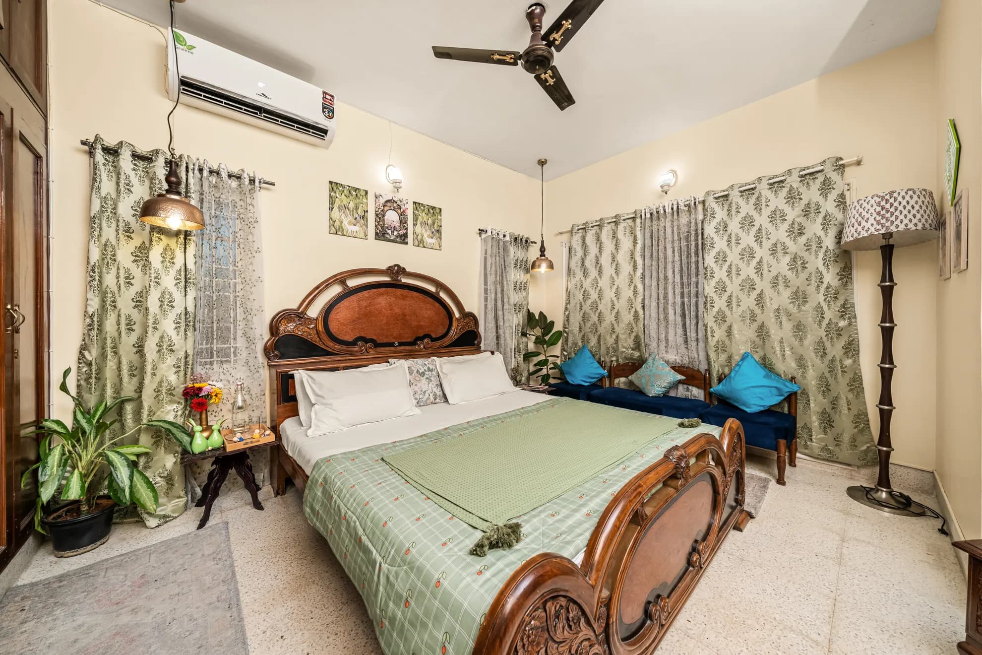 Bedroom with carved wooden double bed, green bedding, ceiling fan, wall-mounted air conditioner and blue velvet bench seating.