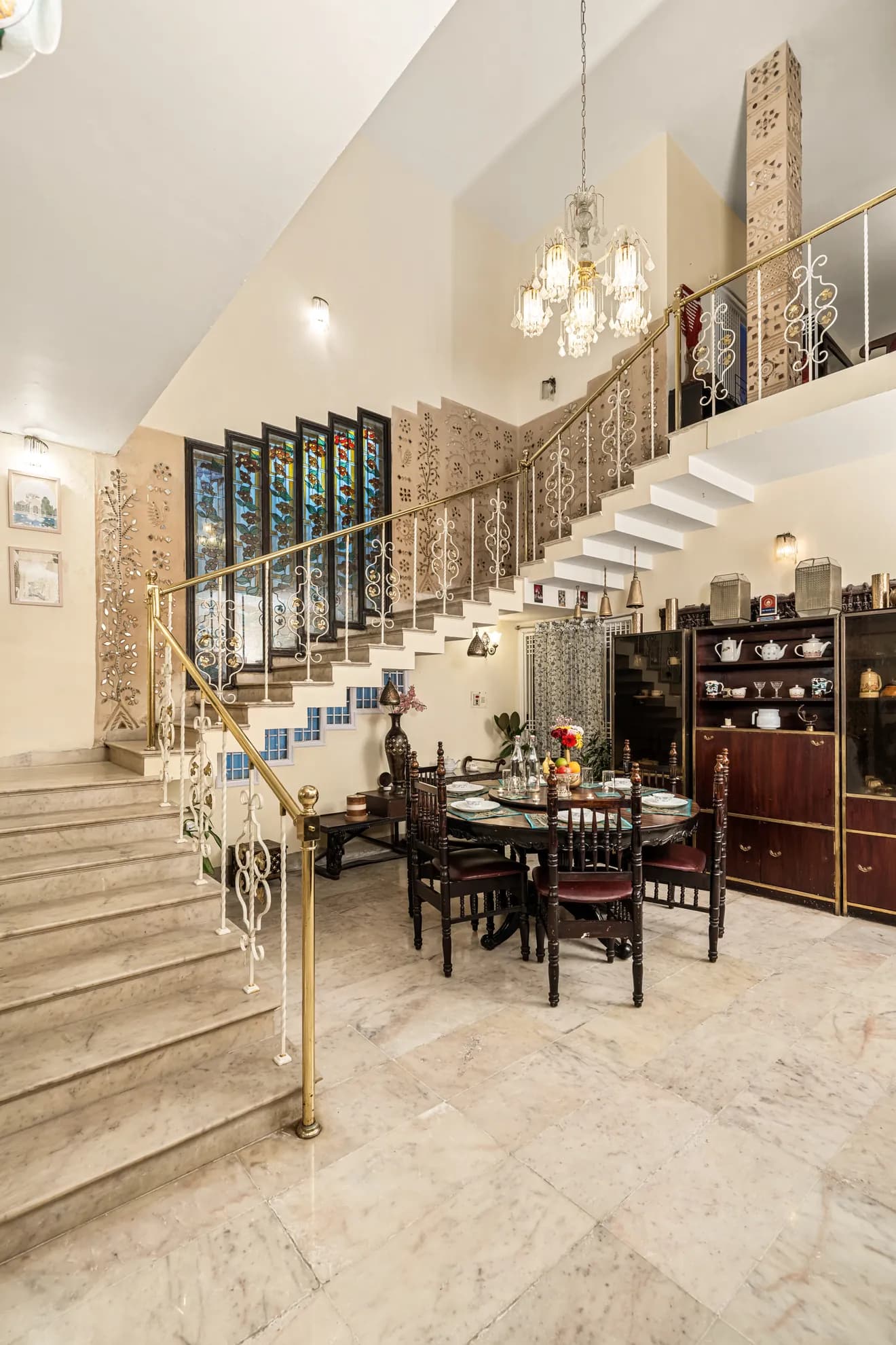 Dining area with a dark wood table and chairs under a chandelier, beside a marble staircase and stained glass windows.