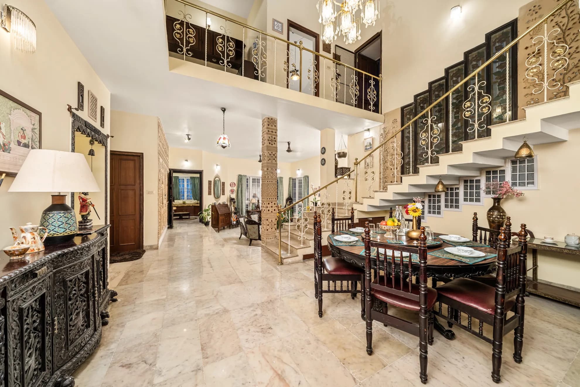 Open double-height dining area with a round wooden table for six, marble floors and an ornate staircase.