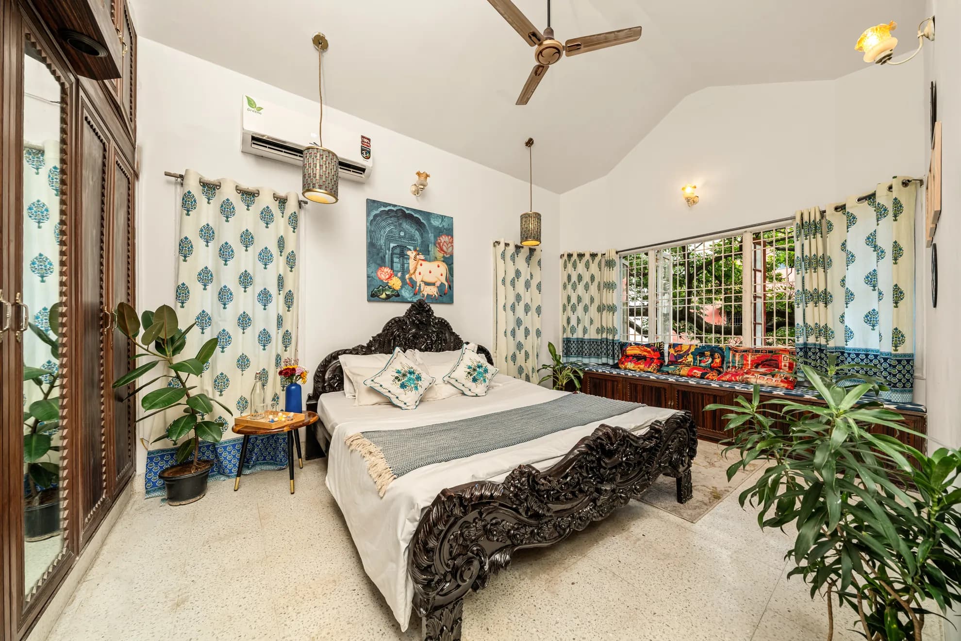 Bedroom with a carved dark wood bed, wall-mounted air conditioner, and a cushioned window seat under leafy garden views.