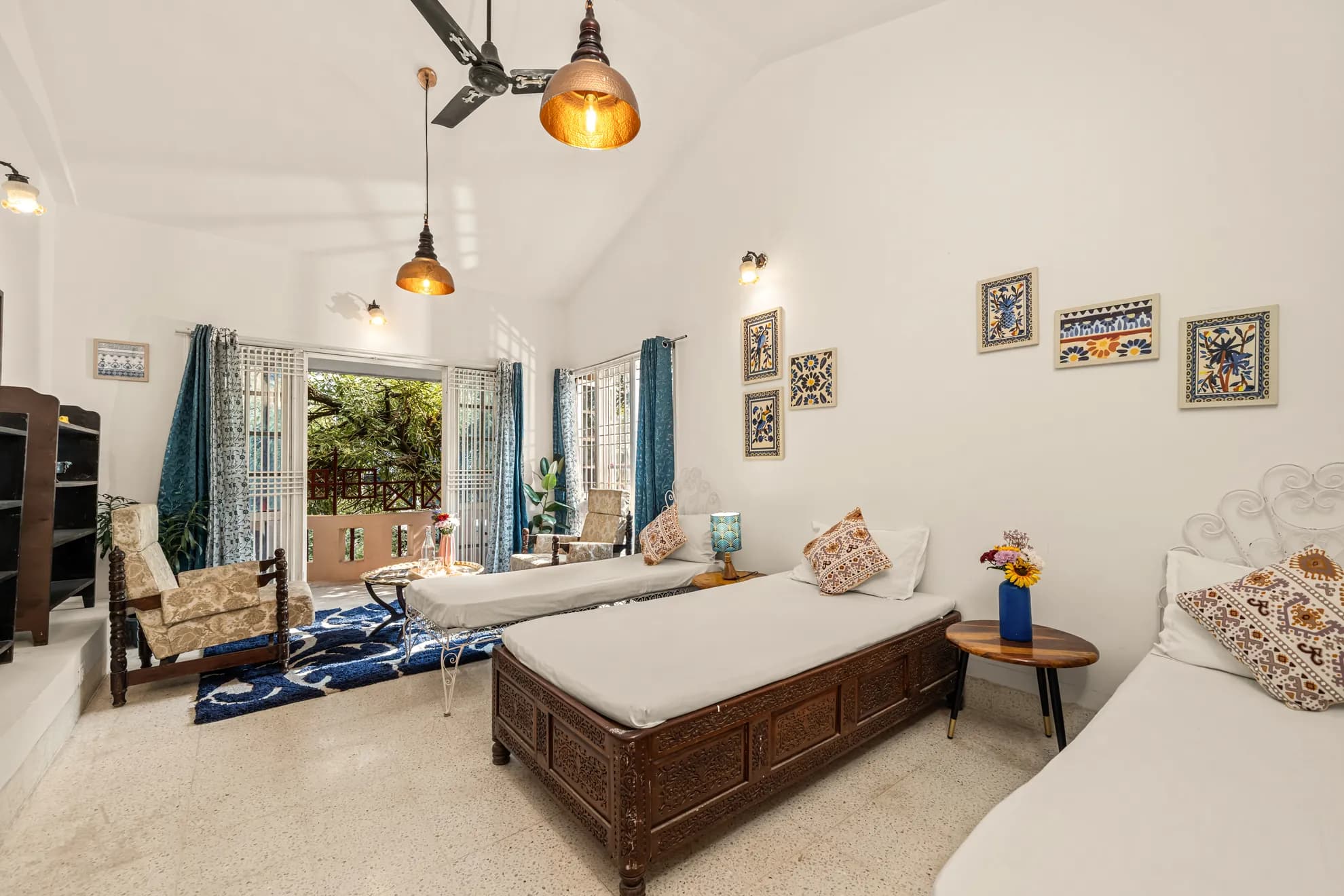 Bedroom with two wooden-framed beds, blue patterned rug, tile wall art and a balcony with greenery beyond.