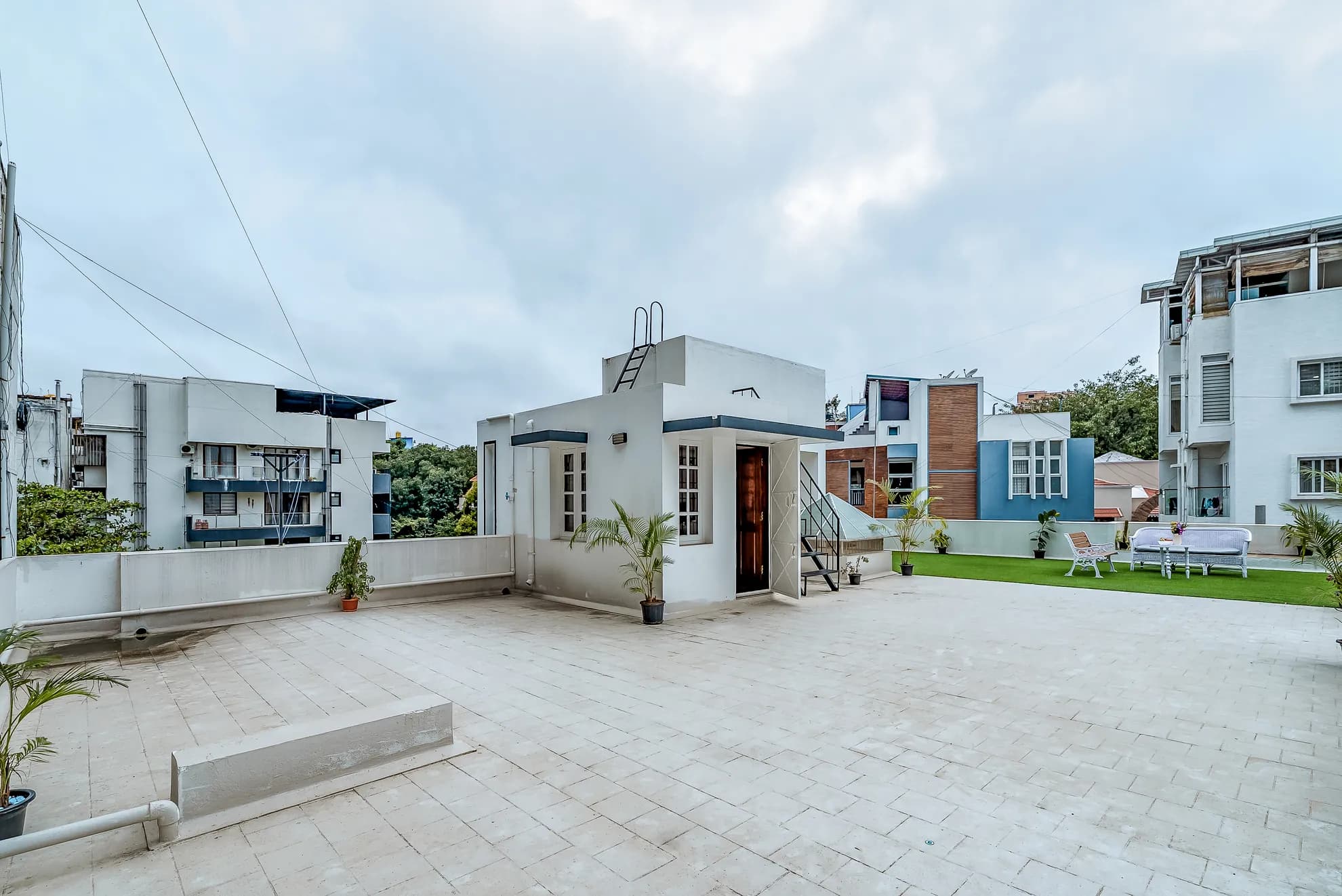 Open paved rooftop terrace with a small white stair-access room, potted palms, and a lawn patch with outdoor seating.