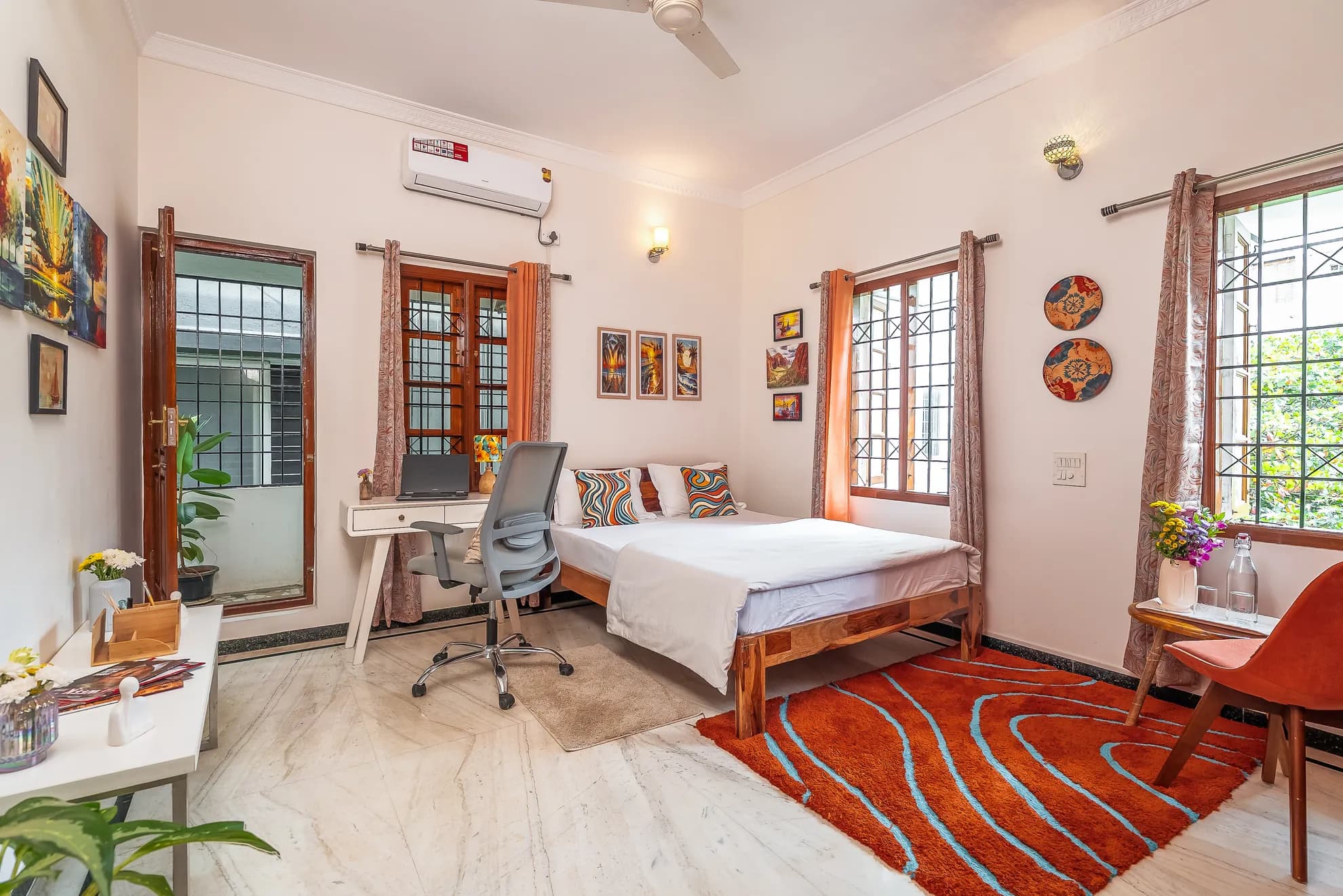 Bedroom with a double bed, wall-mounted air conditioner, a white desk with laptop, and a bold orange rug.