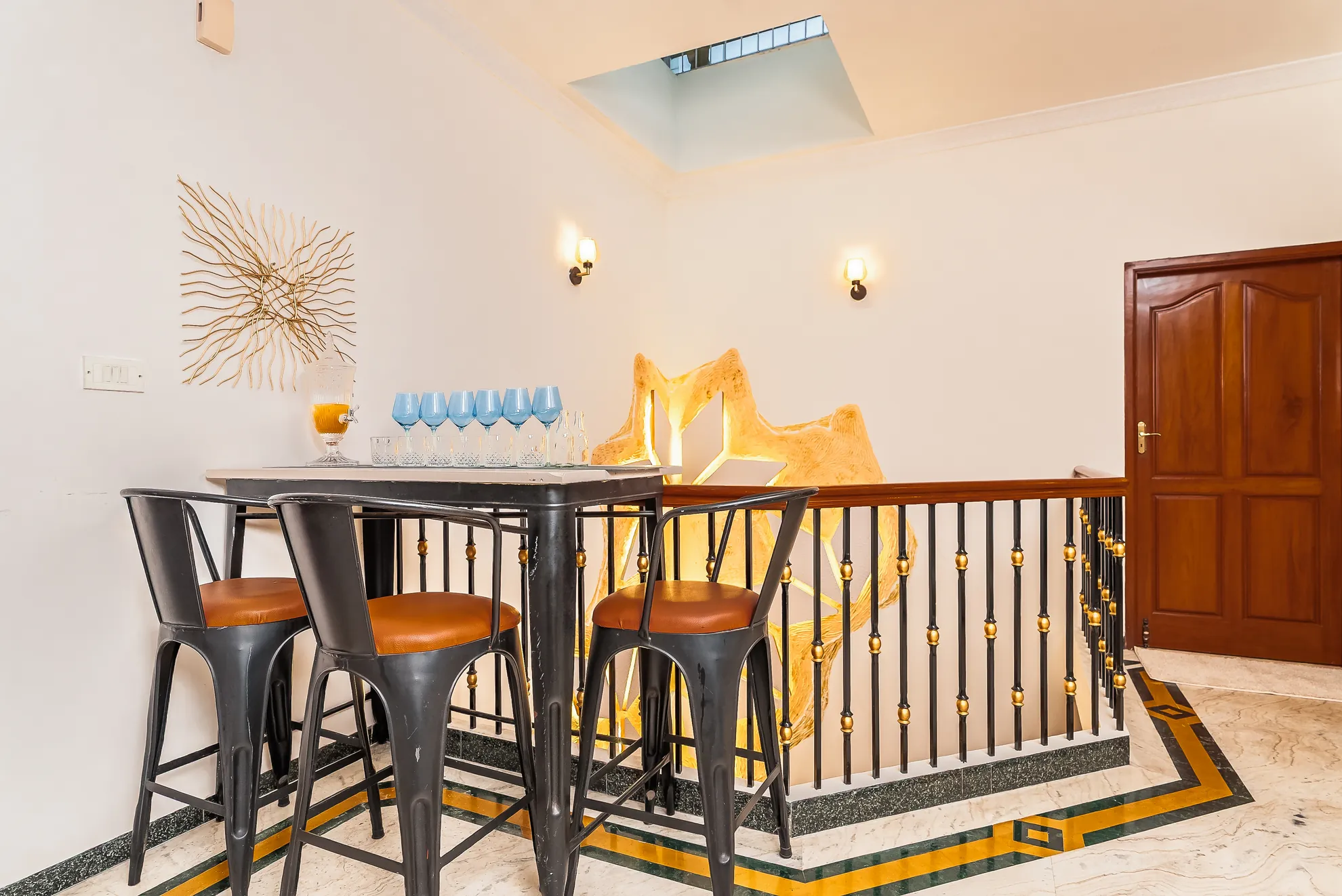 Bar area with a high table, four metal stools and blue wine glasses, beside a wooden railing and gold star decor.