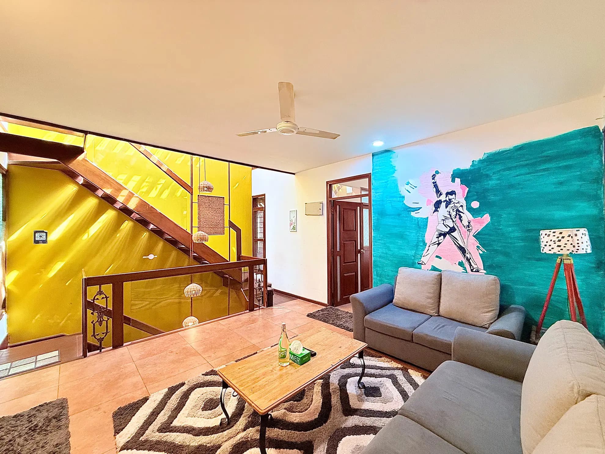 Living room with a yellow staircase wall, teal feature wall with a painted figure, and grey sofas on a patterned rug.