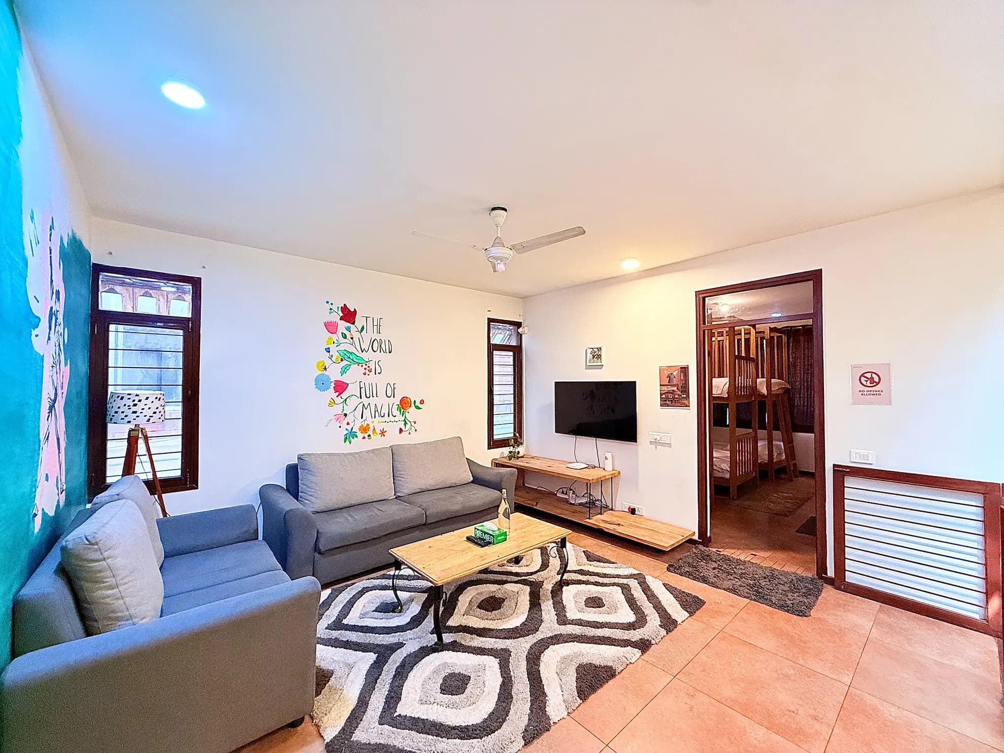 Living room with grey sofas, a wooden coffee table, wall TV and a patterned rug, with bunk beds visible through a doorway.