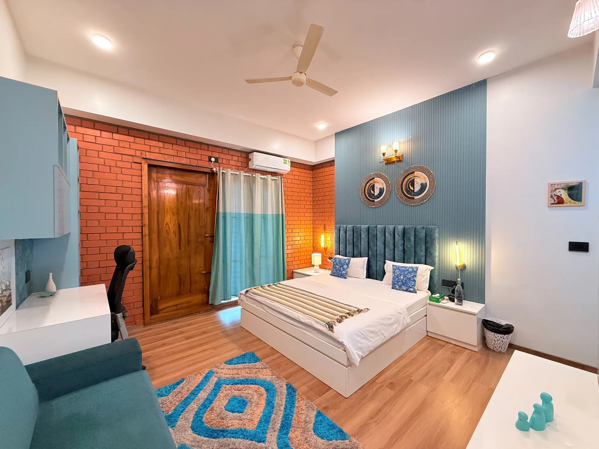 Bedroom with a double bed, exposed brick wall, and teal upholstered headboard beside a wooden door.