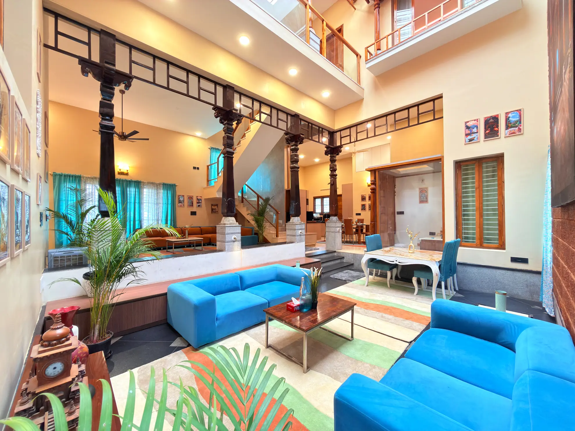 Double-height open living room with blue sofas, a wooden coffee table, and a dining set beyond carved wooden columns.