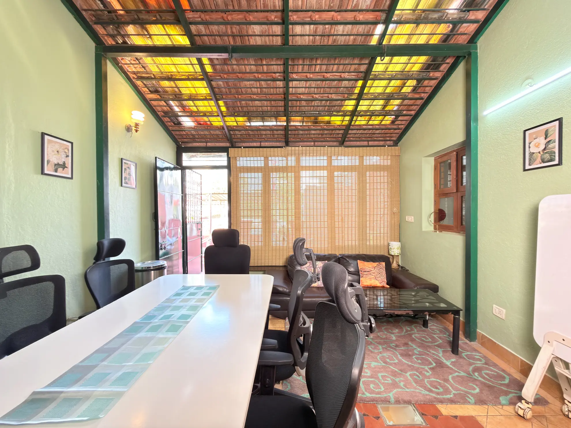 Covered workspace with a long white table, black mesh chairs, and a lounge sofa under a wooden slatted roof.