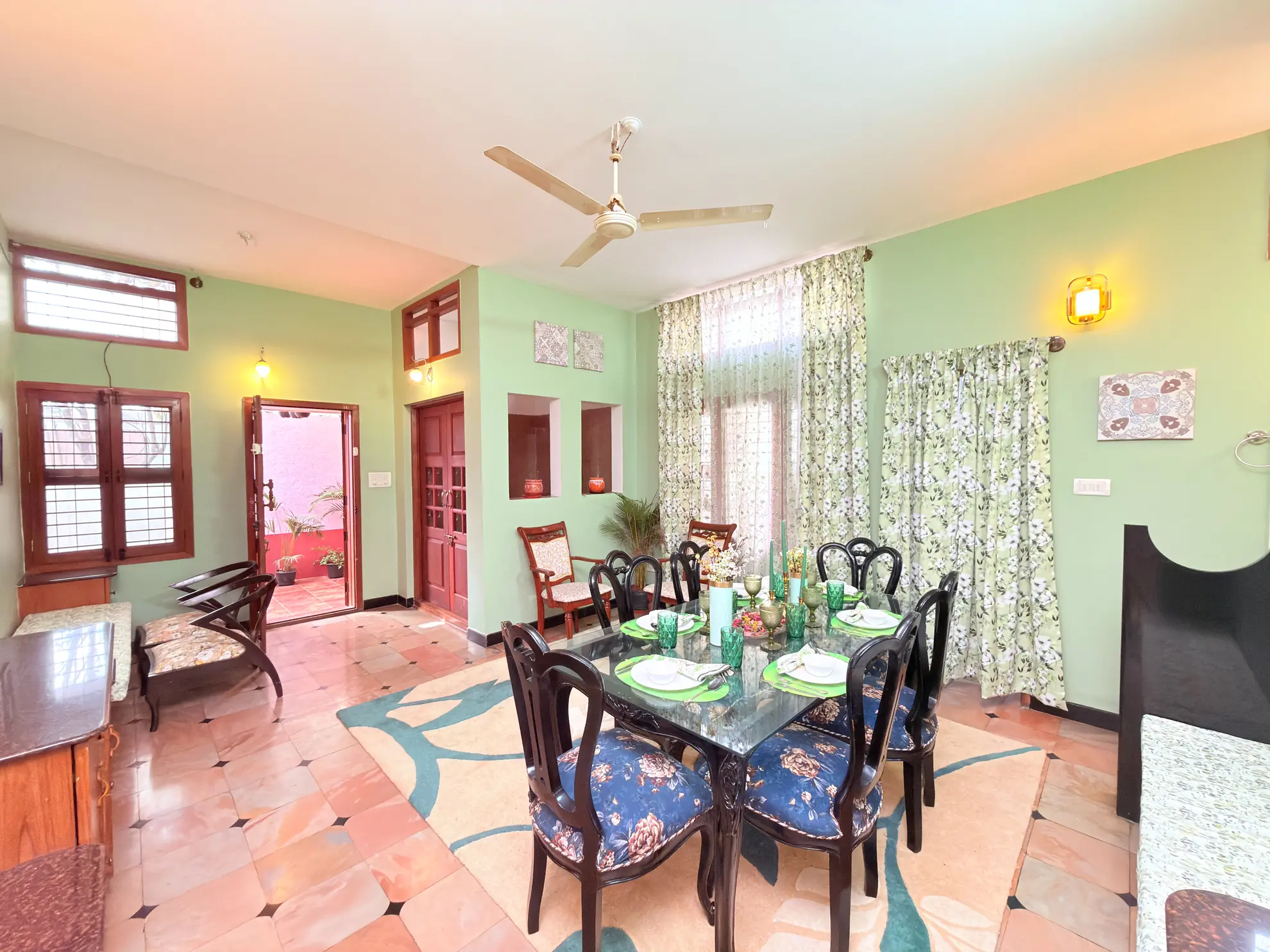 Dining area with a glass table set for four under a ceiling fan, with green walls and a patterned rug.