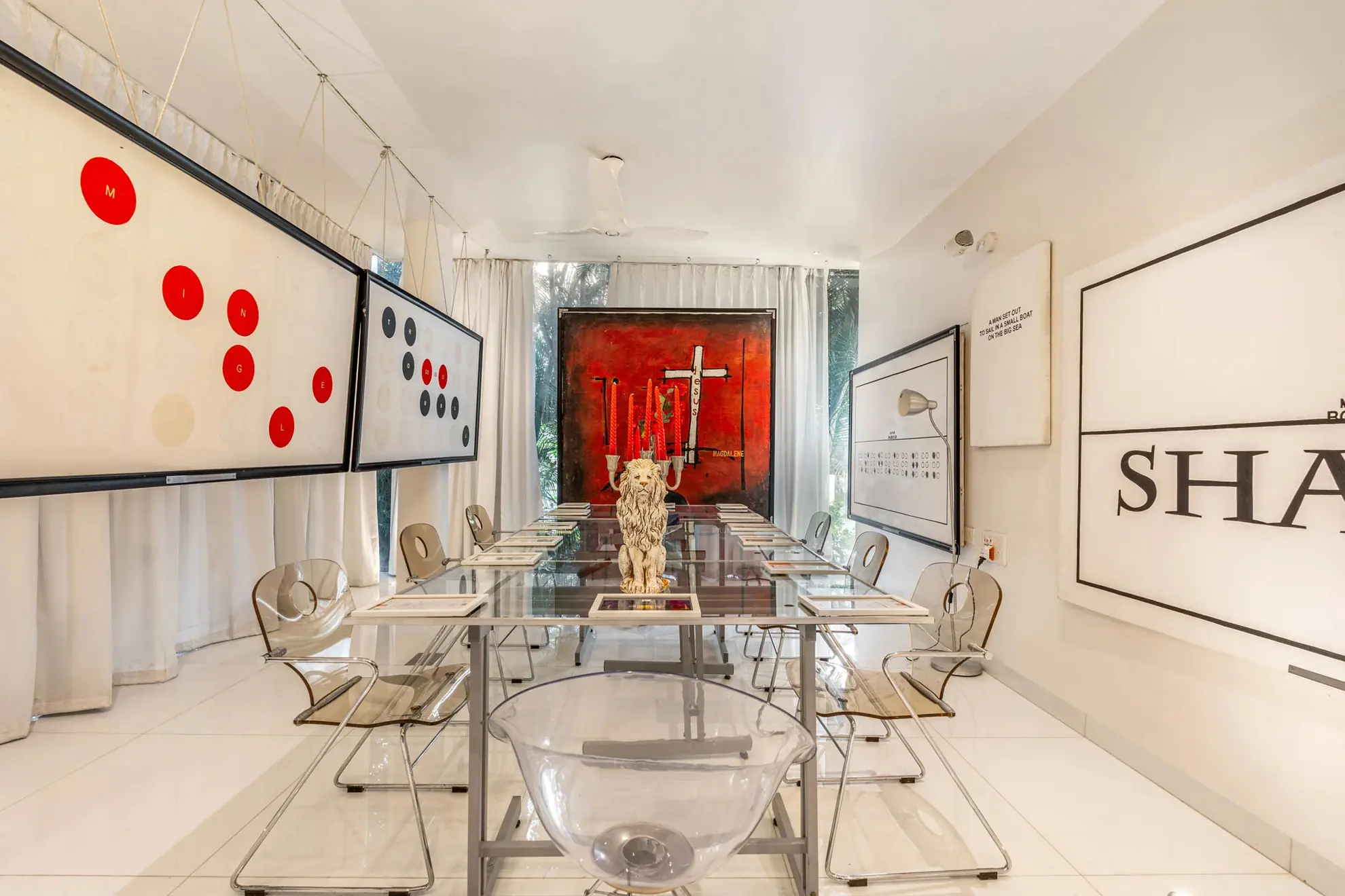 Long glass dining table with clear chairs beside large wall screens and a red painting in a white room.