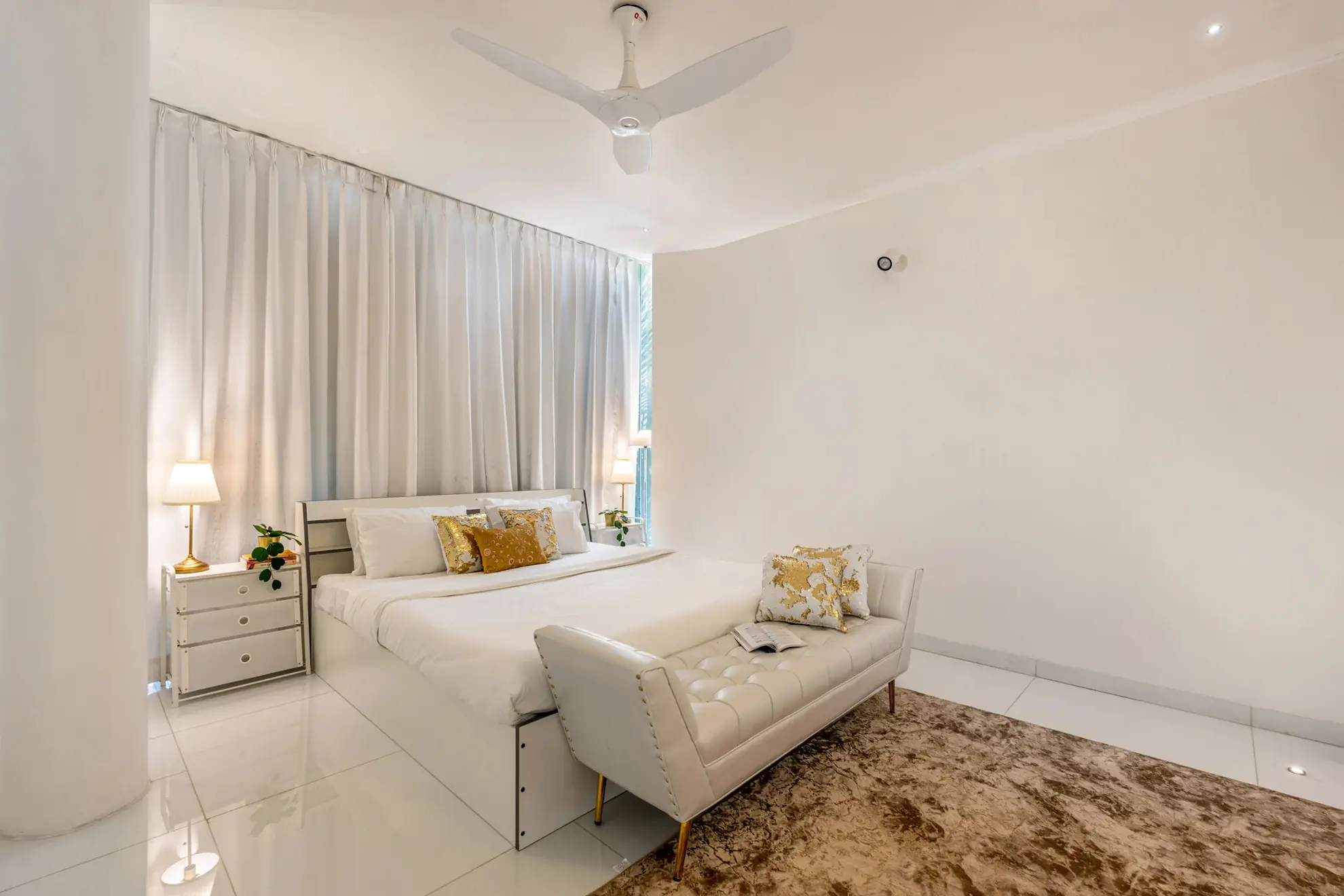 White bedroom with a king bed, gold cushions, a tufted bench and a ceiling fan, lit by bedside lamps.