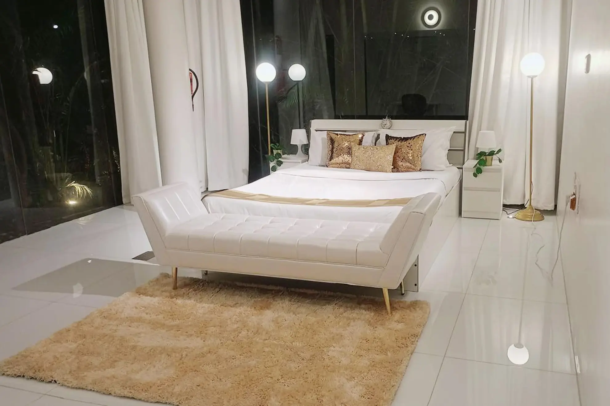 Bedroom with white upholstered bench at foot of bed, gold floor lamps, and shaggy beige rug on white tiles.