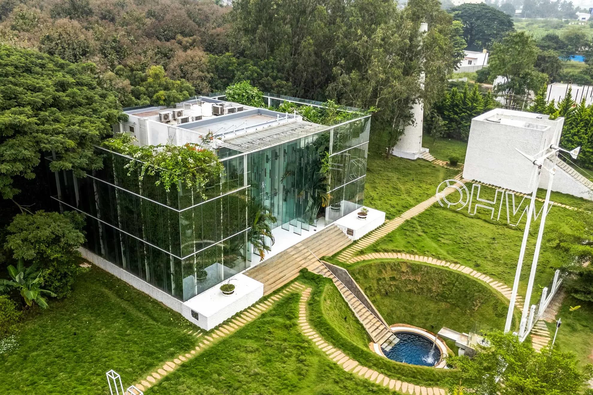 Aerial view of a glass-walled house with a green rooftop, a round sunken pool and stone pathways across a lawn.