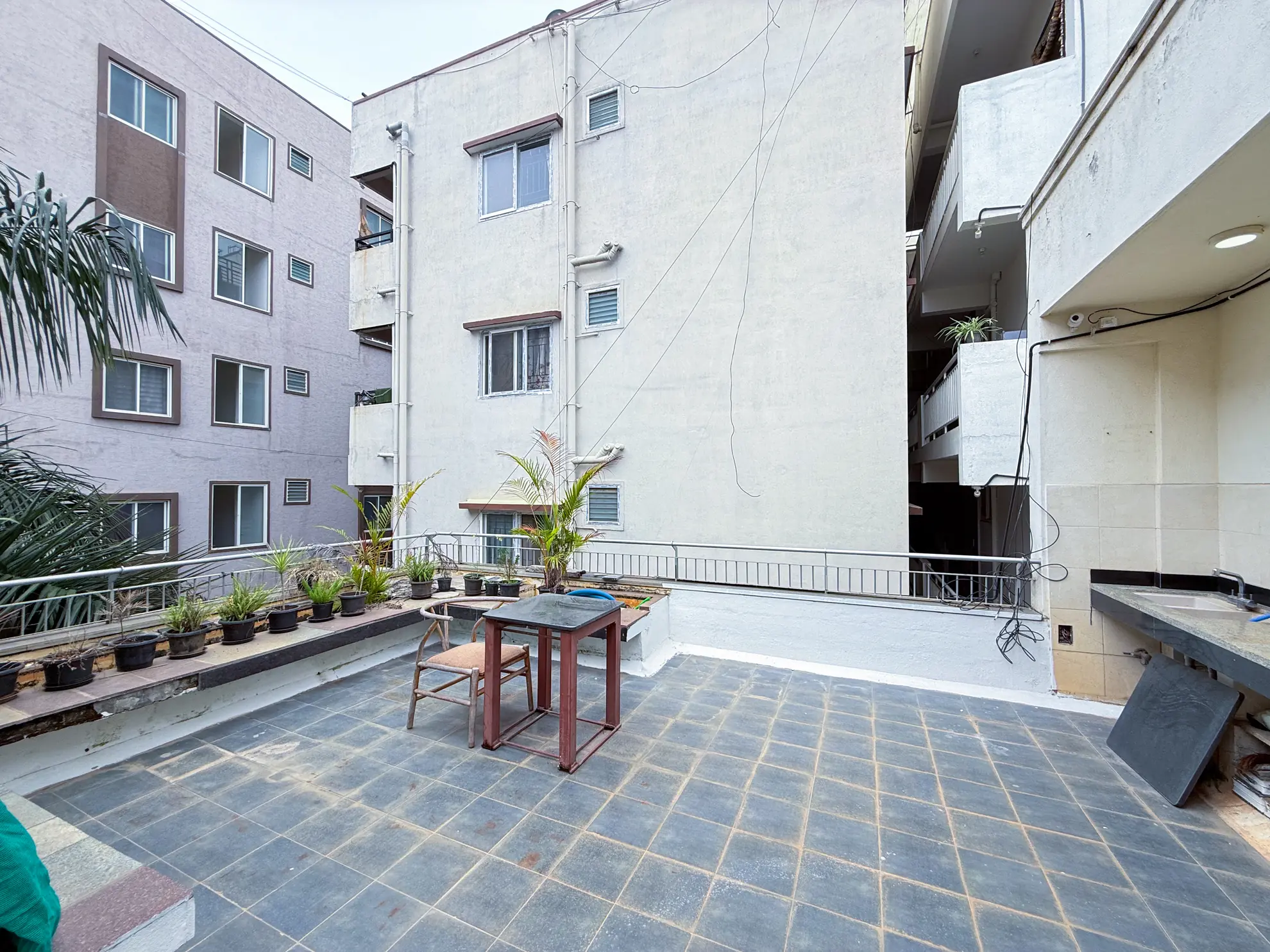 Open tiled terrace with a small table and chair, potted plants along the railing, and an outdoor sink against the wall.