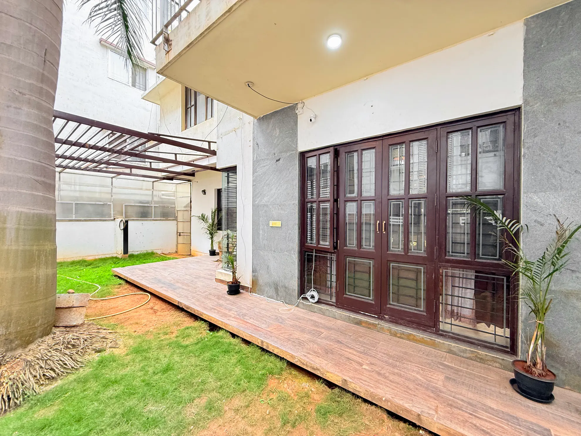 Covered wooden deck beside a garden lawn, with dark-framed glass doors and potted palms outside the house.
