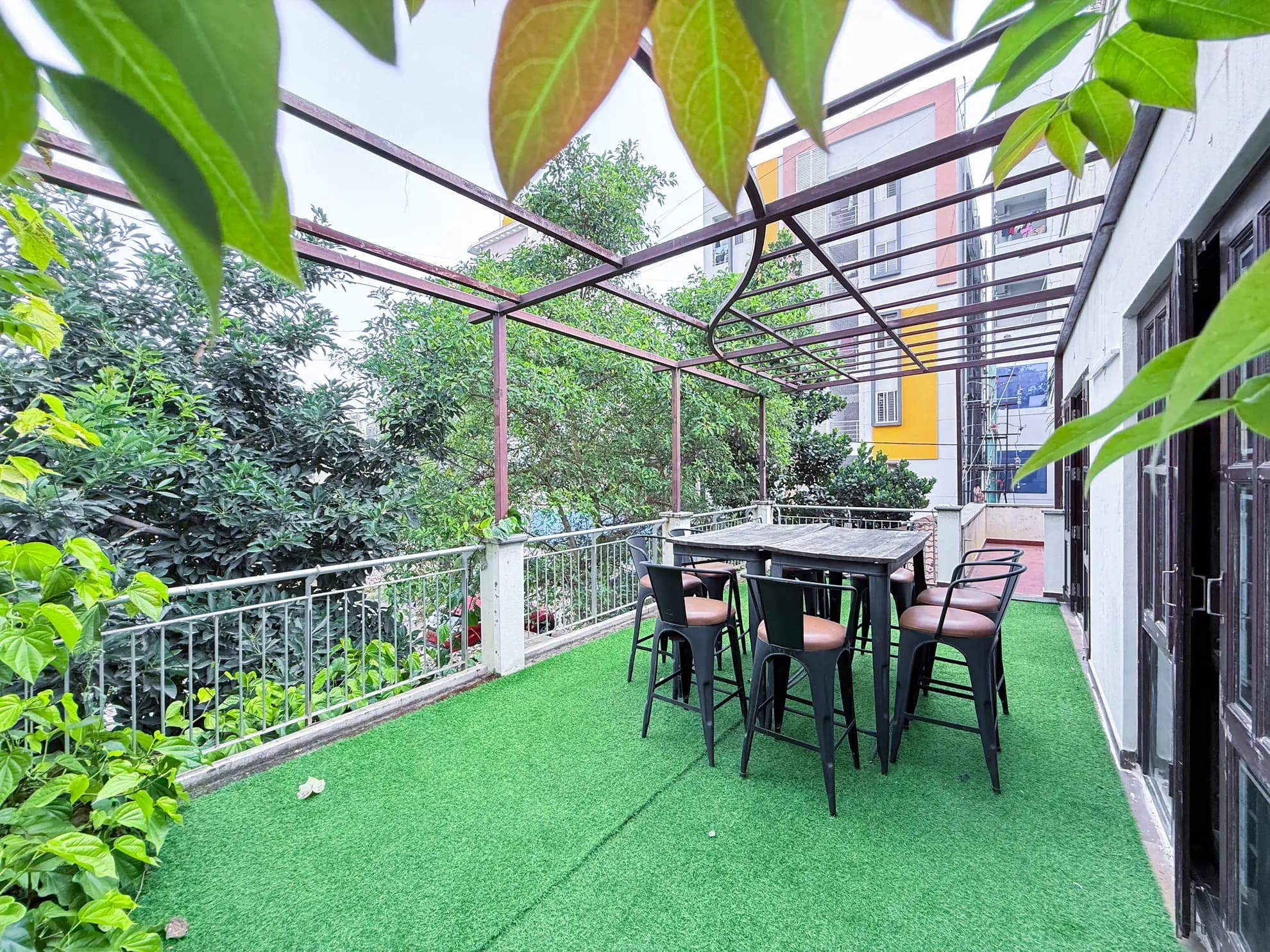 Covered terrace with artificial turf, a wooden high-top table and six black stools under a metal pergola.