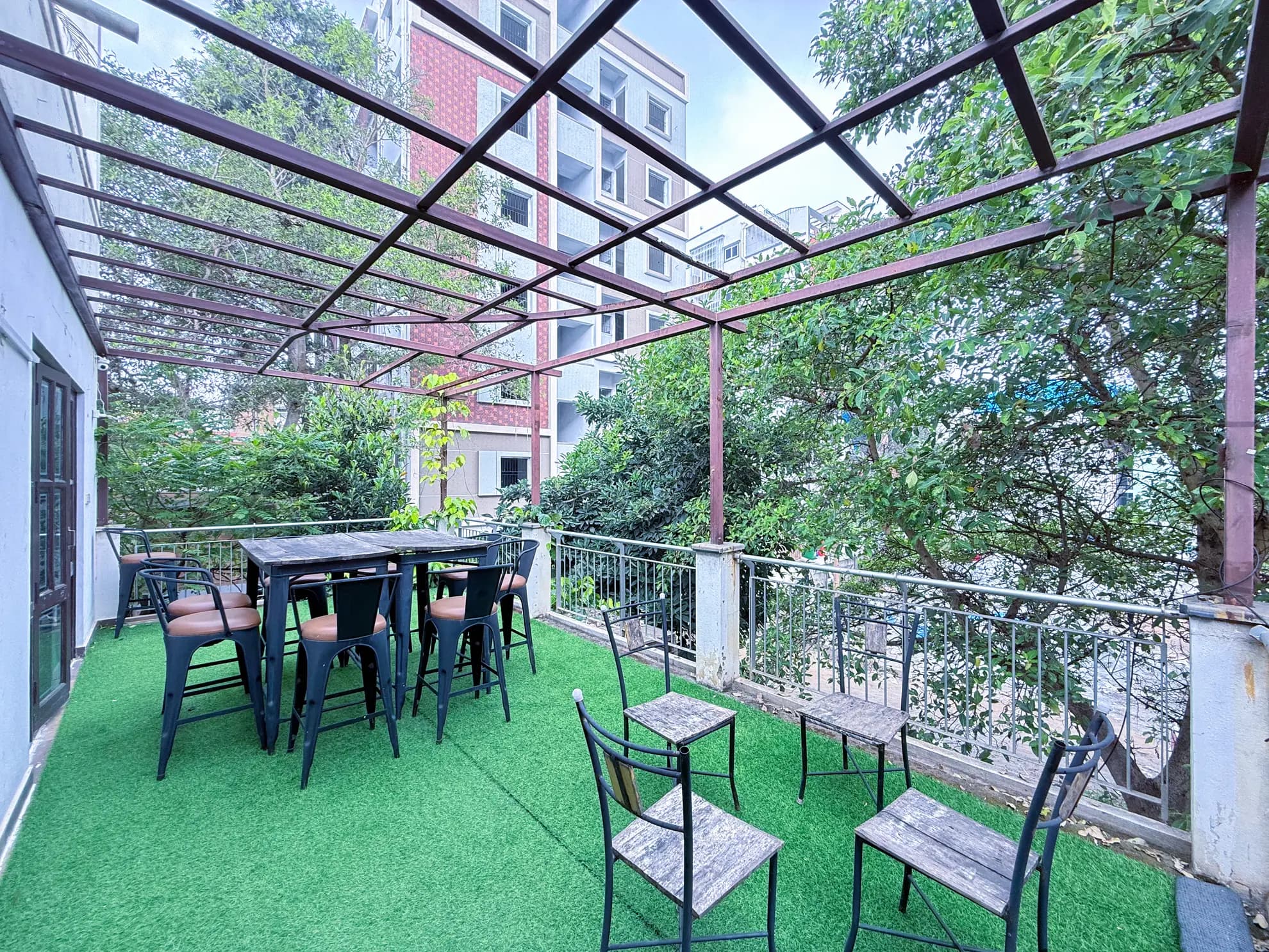 Covered outdoor terrace with artificial grass, a metal pergola, high bar tables and chairs, and trees beyond the railing.